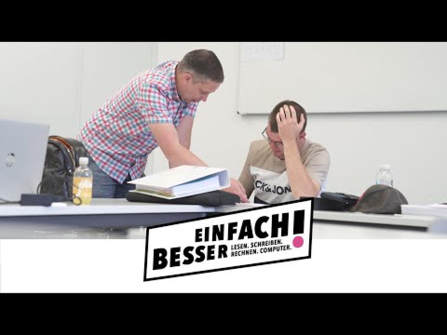Zwei Personen sitzen an einem Schreibtisch mit Papieren und Flaschen; eine steht und zeigt auf ein Dokument, während die andere den Kopf in die Hand stützt. Die Textüberlagerung lautet Einfach! Besser. Lesen. Schreiben. Rechnen. Computer.