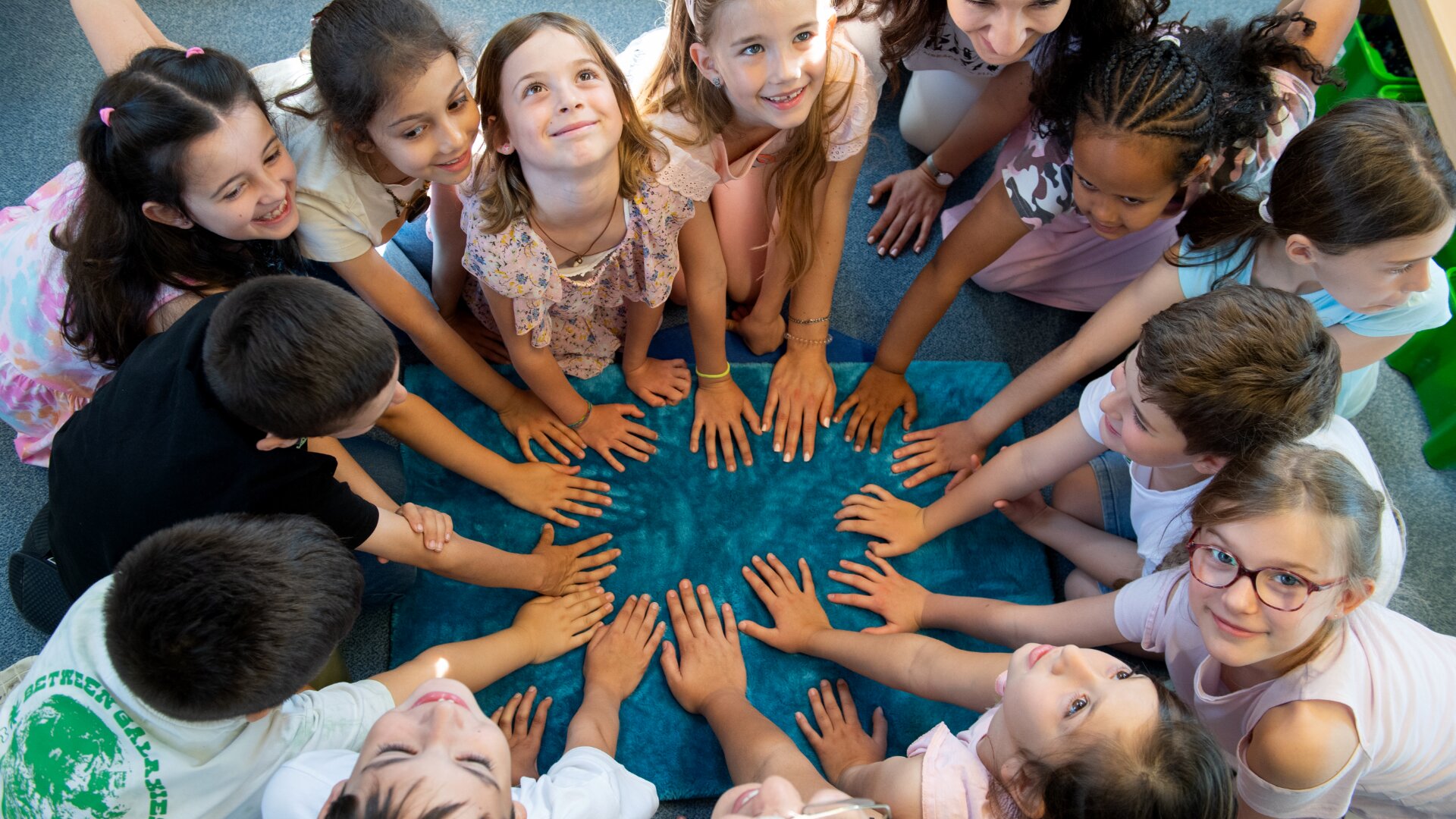 Eine Gruppe von zwölf lächelnden Kindern sitzt in einem Kreis, legt ihre Hände auf einer blauen Matte in der Mitte zusammen, blickt auf und wirkt glücklich und engagiert.