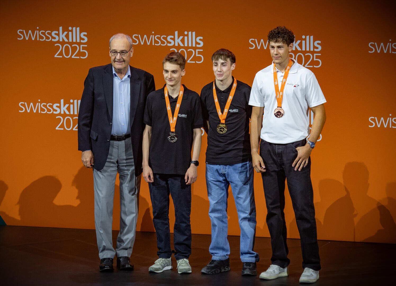 Vier Männer stehen vor einer orangefarbenen Kulisse der SwissSkills 2025; drei junge Männer mit Medaillen und Freizeitkleidung stehen neben einem älteren Mann im Anzug, alle lächeln für ein Gruppenfoto.