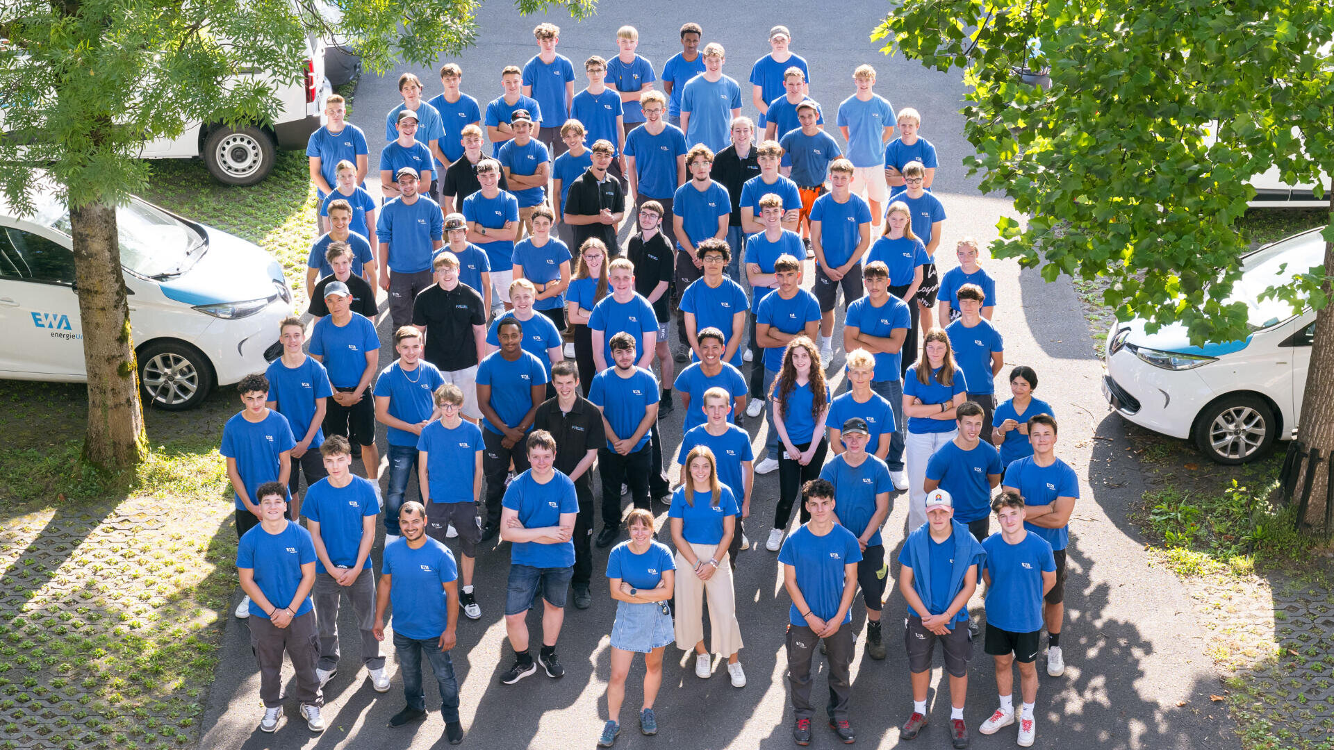 Eine große Gruppe von Menschen in blauen Hemden steht an einem sonnigen Tag im Freien in einer halb organisierten Formation, lächelnd und der Kamera zugewandt, umgeben von Bäumen und geparkten weißen Autos.