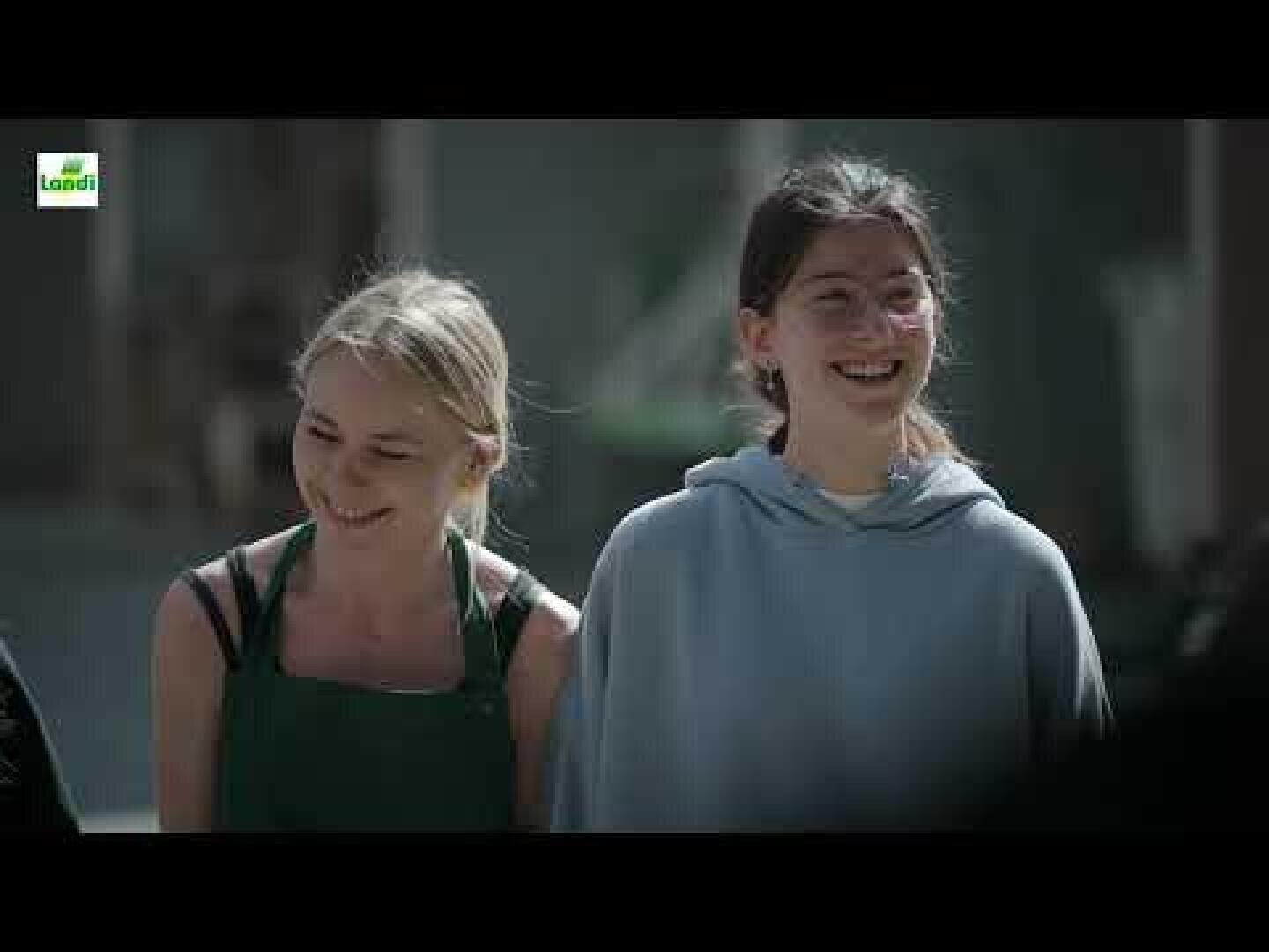 Zwei junge Frauen stehen im Freien nebeneinander, lächeln und schauen nach rechts. Die eine trägt eine grüne Schürze über einem schwarzen Oberteil, die andere eine Brille und einen hellblauen Kapuzenpullover. Der Hintergrund ist unscharf.