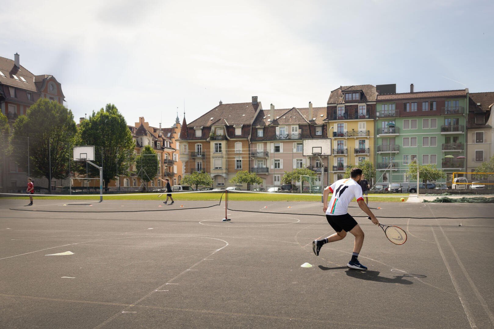 Zwei Personen spielen Tennis auf einem Außenplatz in einem städtischen Gebiet, mit bunten Wohngebäuden und Basketballkörben im Hintergrund unter einem sonnigen Himmel.