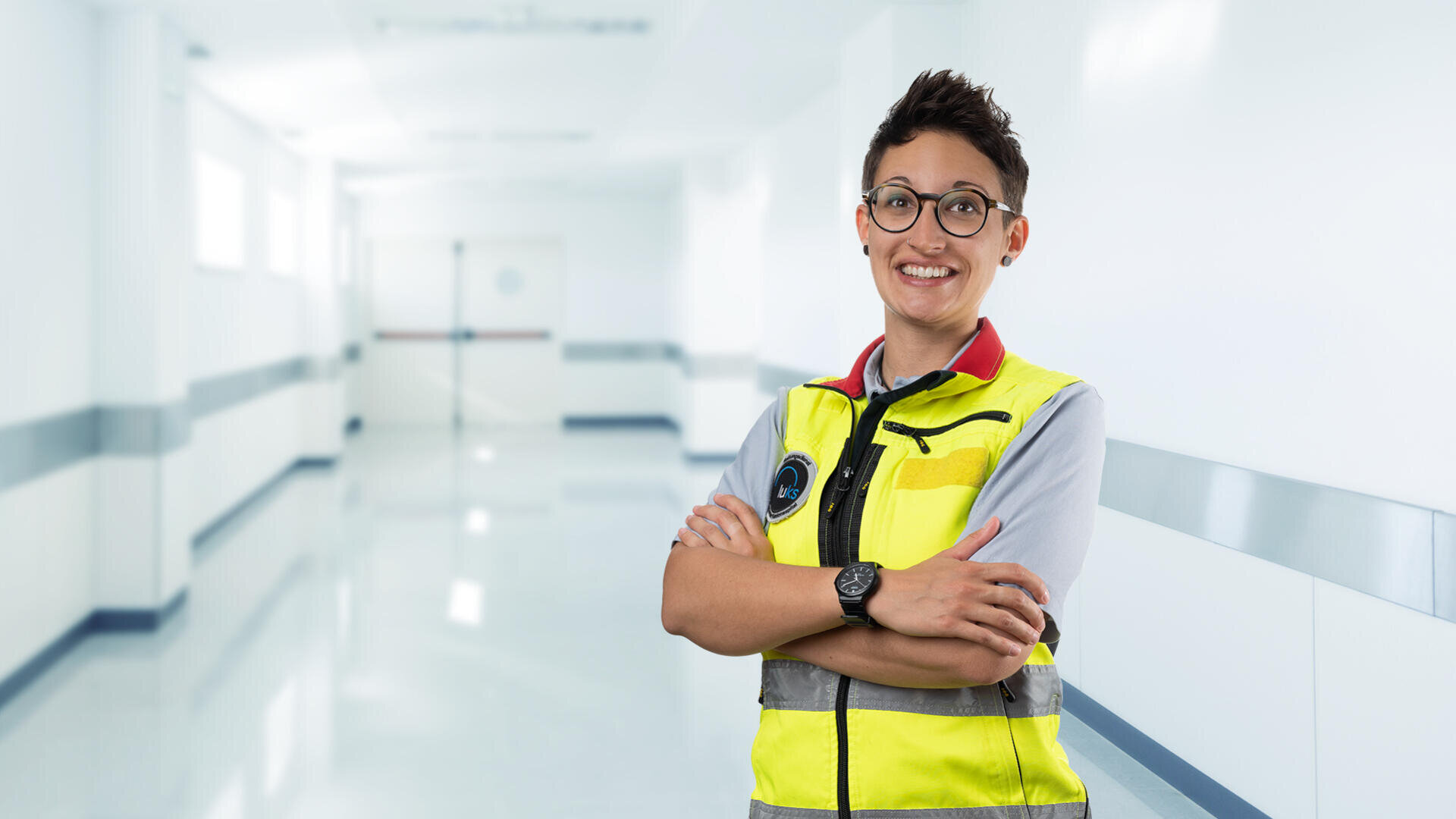 Eine lächelnde Person mit Brille und gelber Sicherheitsweste steht mit verschränkten Armen in einem hellen, modernen Flur, wahrscheinlich in einem Krankenhaus oder einer medizinischen Einrichtung.