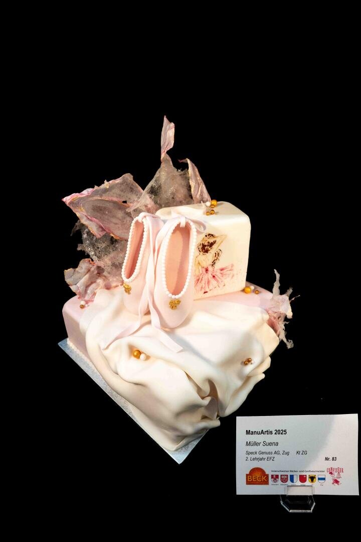 Eine elegante Torte, verziert mit Ballett-Pantoffeln aus Fondant, zarten rosa und weißen Rüschen sowie goldenen Akzenten. Sie ist mit einem aufgedruckten Bild einer Ballerina und Zuckerdekorationen verziert. Vorne ist eine Glückwunschkarte angebracht.