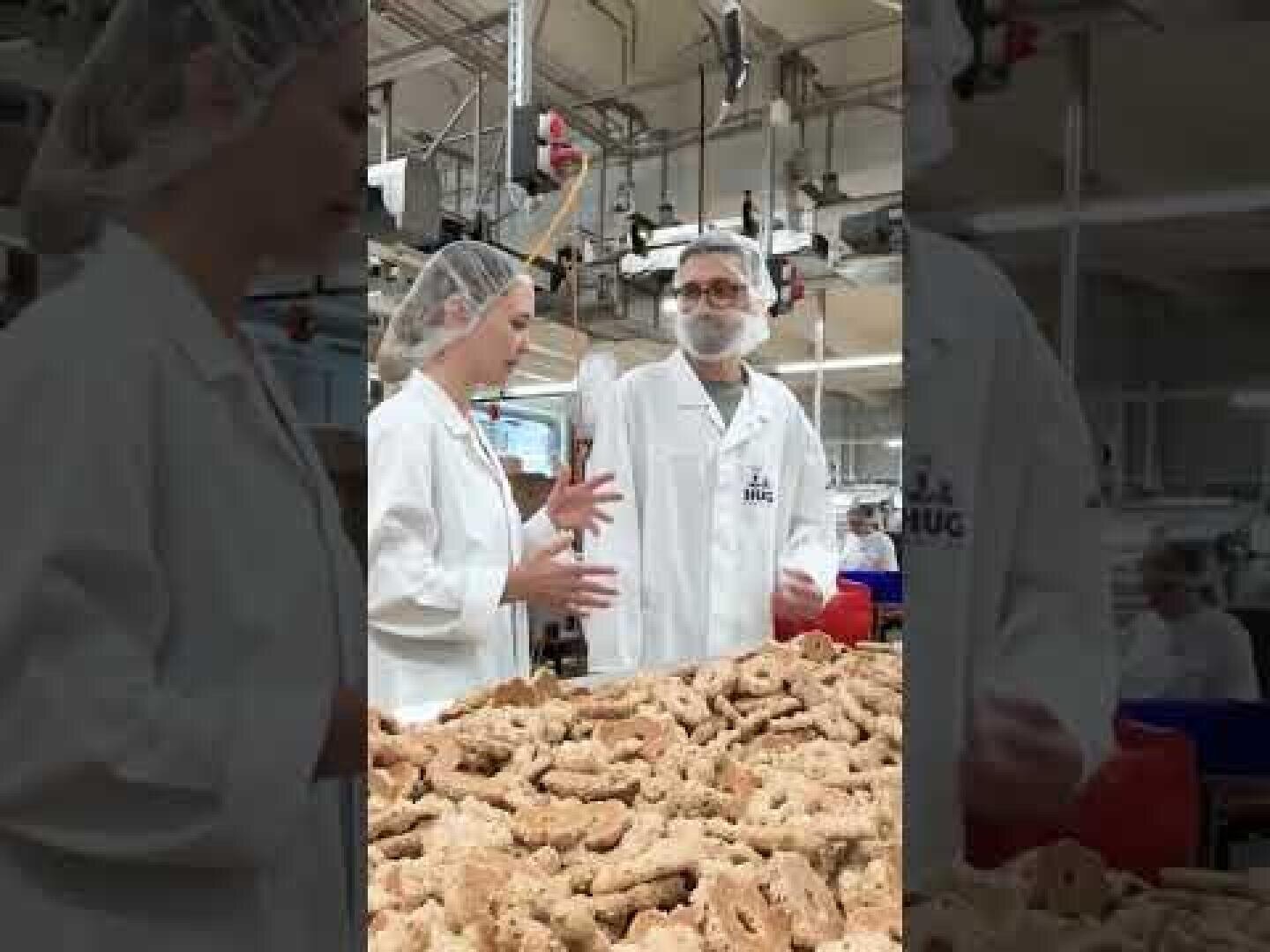 Zwei Personen in weißen Laborkitteln und Haarnetzen unterhalten sich in einer Fabrikumgebung. Sie stehen hinter einem großen Stapel von Leckereien in Form von Hundeknochen auf einem Tisch, und im Hintergrund sind Industrieanlagen zu sehen.