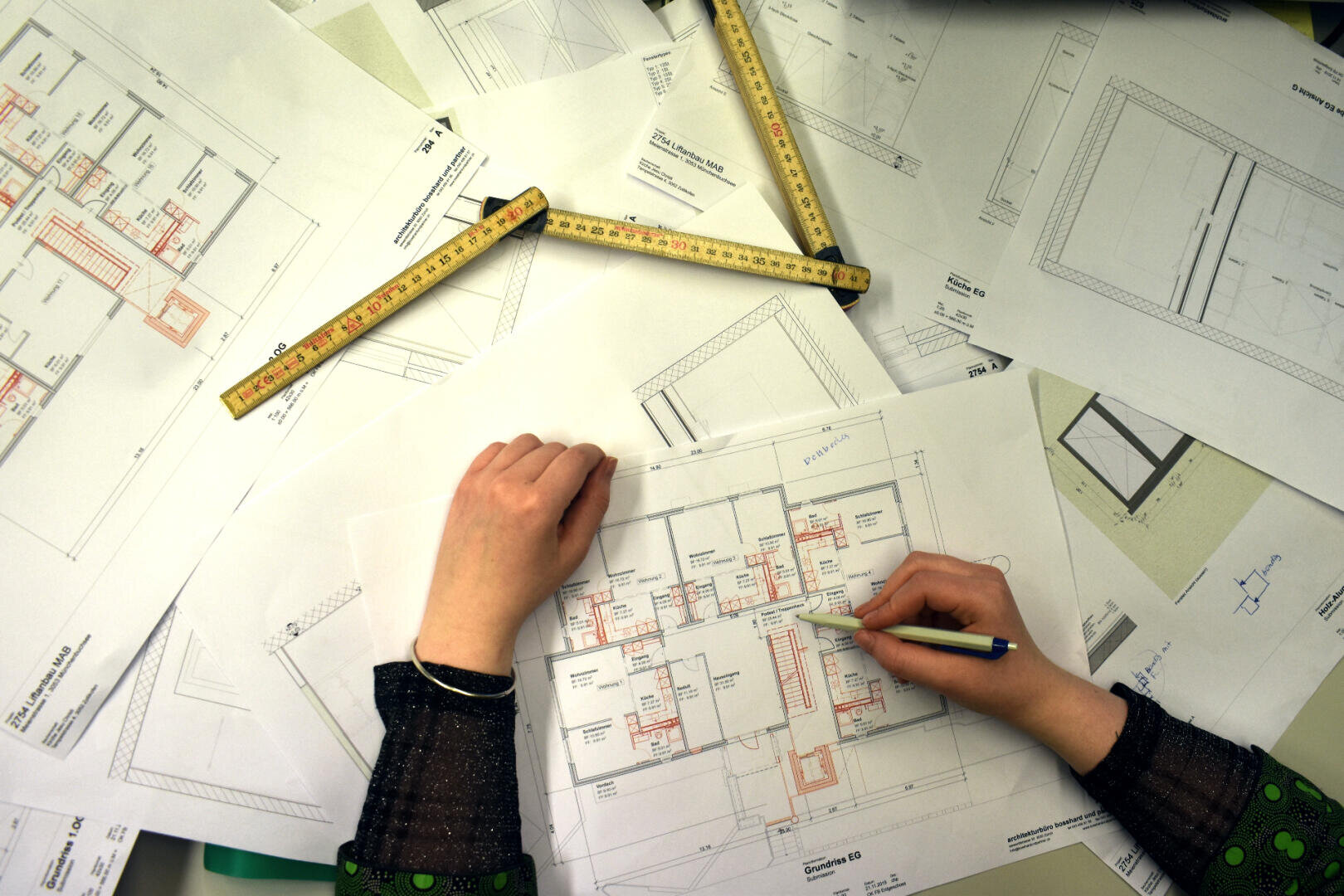 Eine Person hält einen Bleistift in der Hand und zeigt auf einen Gebäudegrundriss, umgeben von verschiedenen Architekturzeichnungen und Zollstöcken, die auf einem Schreibtisch ausgebreitet sind.