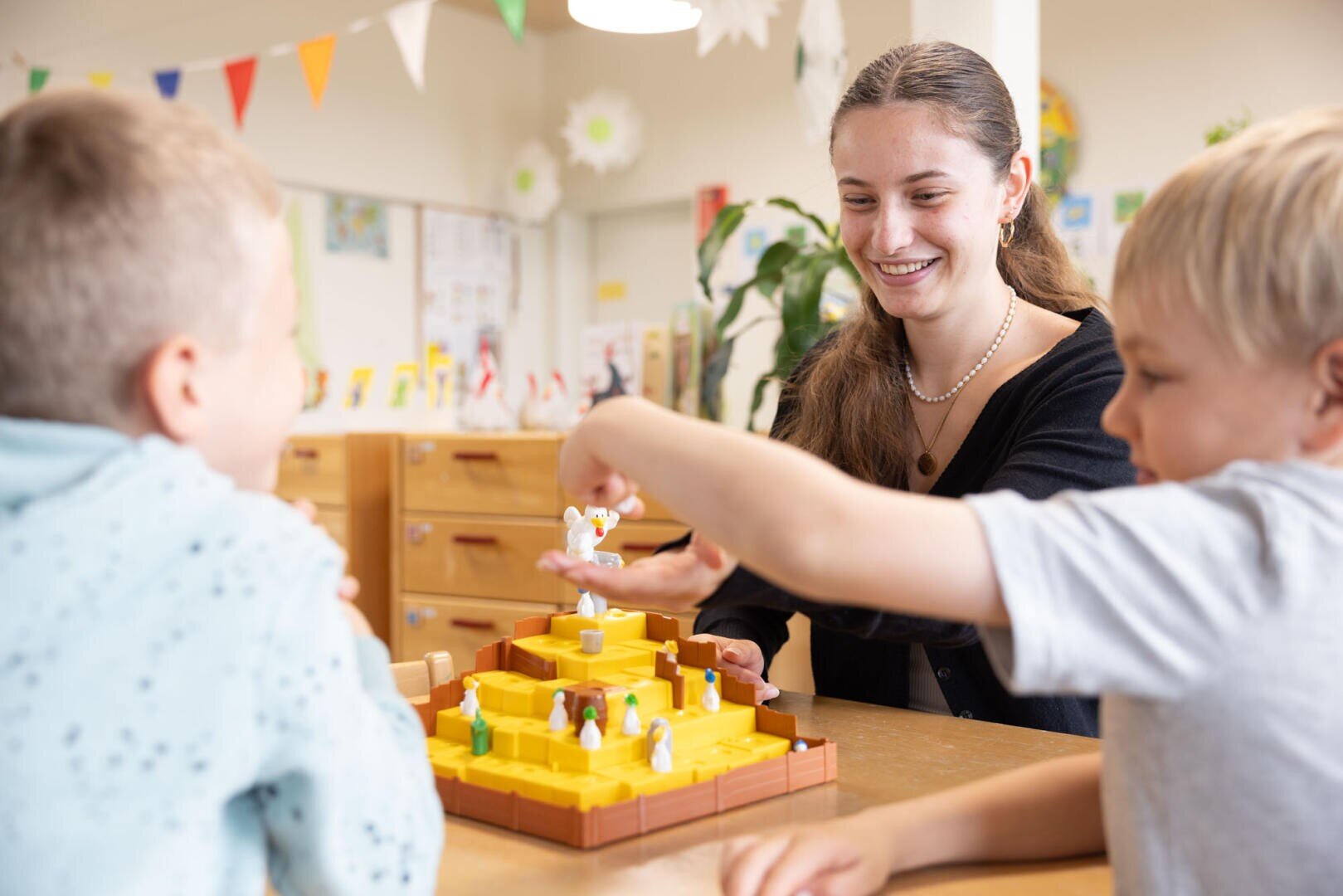 Eine Frau und zwei Jungen sitzen lächelnd an einem Tisch und spielen ein buntes Brettspiel mit kleinen Figuren in einem hellen, dekorierten Klassenzimmer.