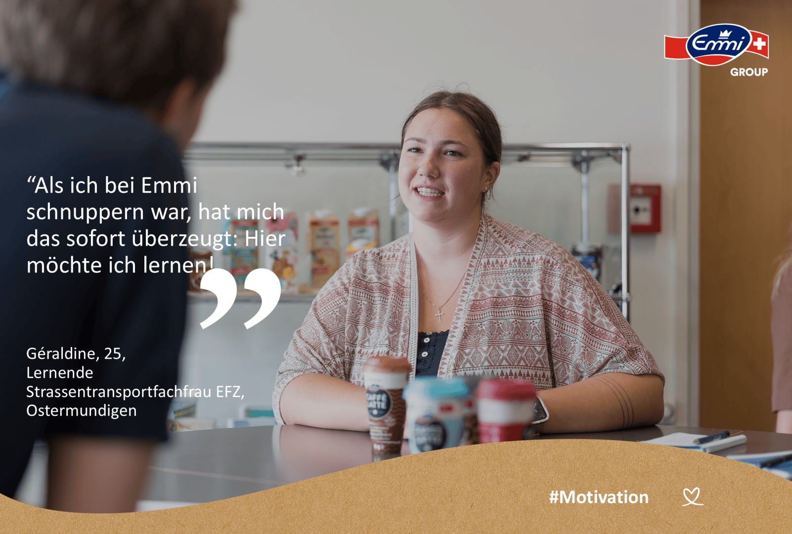 Eine junge Frau sitzt an einem Tisch mit Kaffeetassen und lächelt jemanden aus dem Off an. Der Schriftzug des Unternehmens Emmi ist zu sehen, zusammen mit einem Zitat über die Freude an ihrer Lernerfahrung bei Emmi. Am unteren Rand wird #Motivation angezeigt.