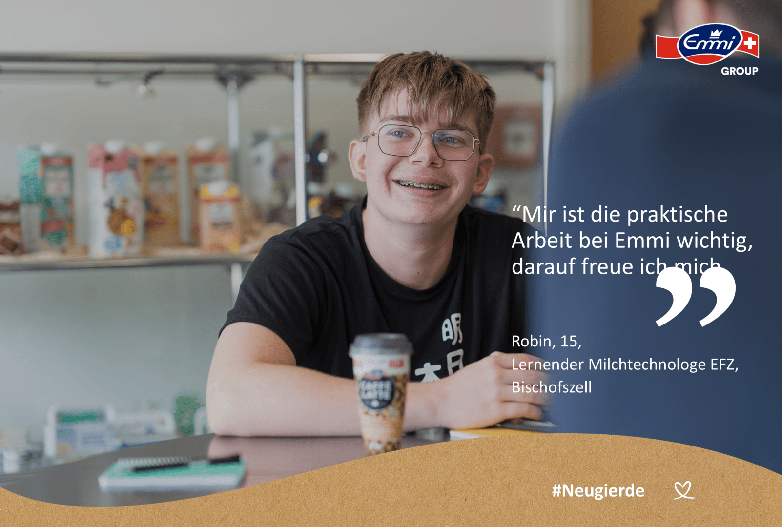 Ein lächelnder Teenager mit Brille sitzt an einem Tisch mit einem Getränk, in einem hellen Raum mit Regalen mit Produkten hinter ihm. Auf dem Bild erscheinen ein deutscher Text und das Emmi-Logo.