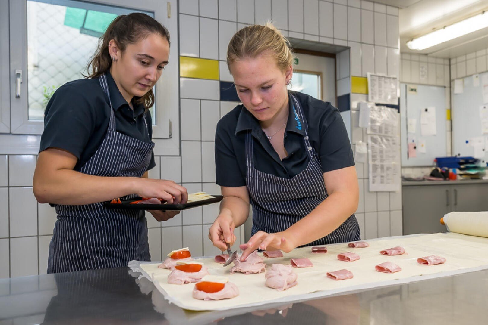 Zwei Mitarbeiter in gestreiften Schürzen bereiten in einer Großküche kleine Gebäckhäppchen zu, die mit Schinken und Tomatenscheiben belegt sind.