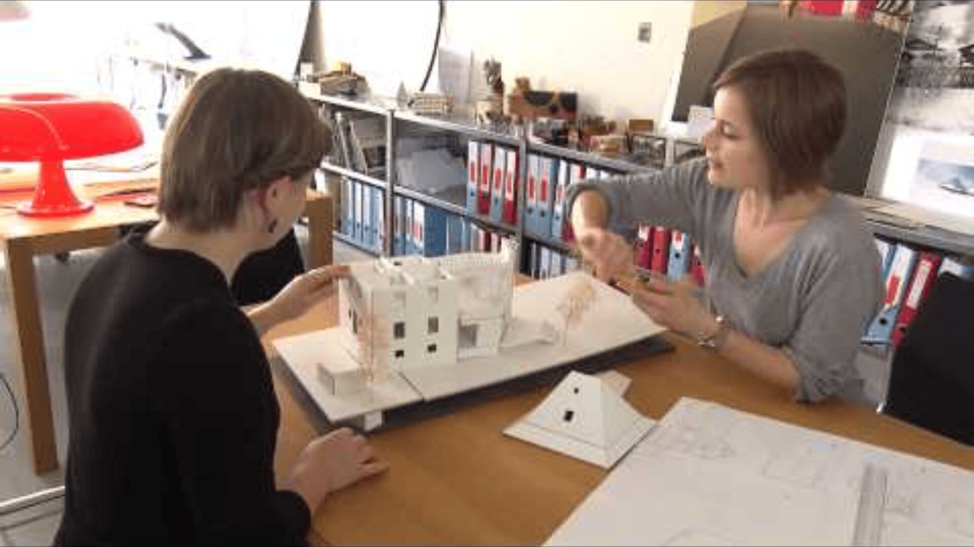 Zwei Frauen sitzen an einem Tisch und diskutieren über ein weißes Architekturmodell eines Gebäudes. Im Hintergrund sind Papierpläne, Regale mit Aktenordnern und eine rote Lampe zu sehen.