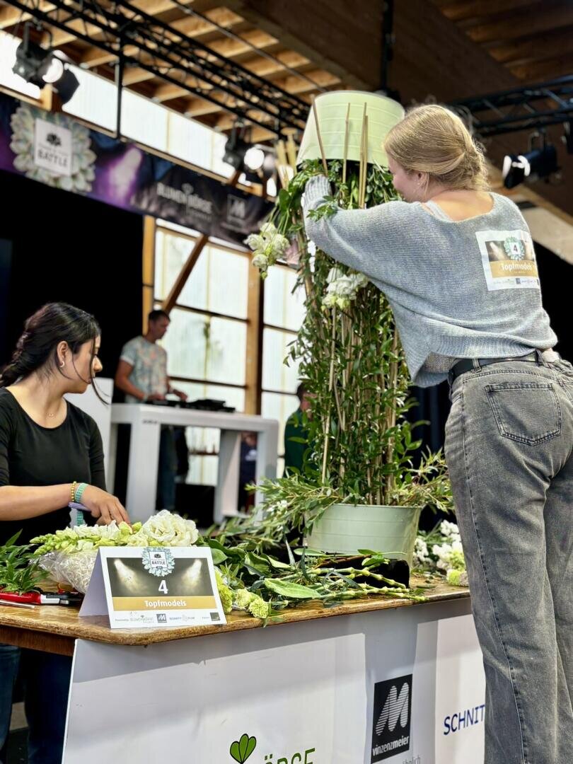 Zwei Personen arrangieren Blumen an einem Wettbewerbstisch; eine steht, um ein hohes Blumenarrangement zu gestalten, während die andere sitzt und Blumen arrangiert. Auf dem Tisch liegen verschiedene Blumen und Grünpflanzen, daneben steht ein Schild mit der Nummer 4.