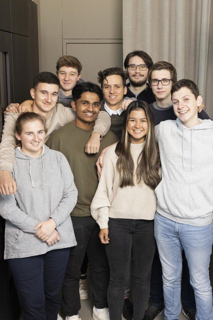 Eine Gruppe von acht jungen Erwachsenen, fünf Männer und drei Frauen, steht in einem Haus eng beieinander und lächelt in die Kamera. Sie sind leger in Pullover und Kapuzenpullover gekleidet und stehen vor einem neutralen Hintergrund.