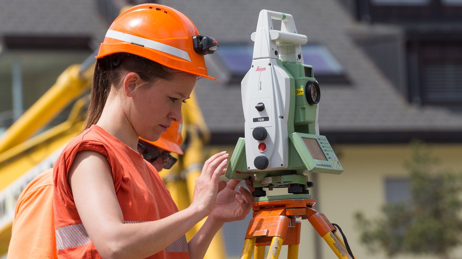 Eine Frau mit orangefarbenem Schutzhelm und Weste bedient ein Vermessungsinstrument auf einem Stativ auf einer Baustelle, mit unscharfen Baumaschinen und Gebäuden im Hintergrund.
