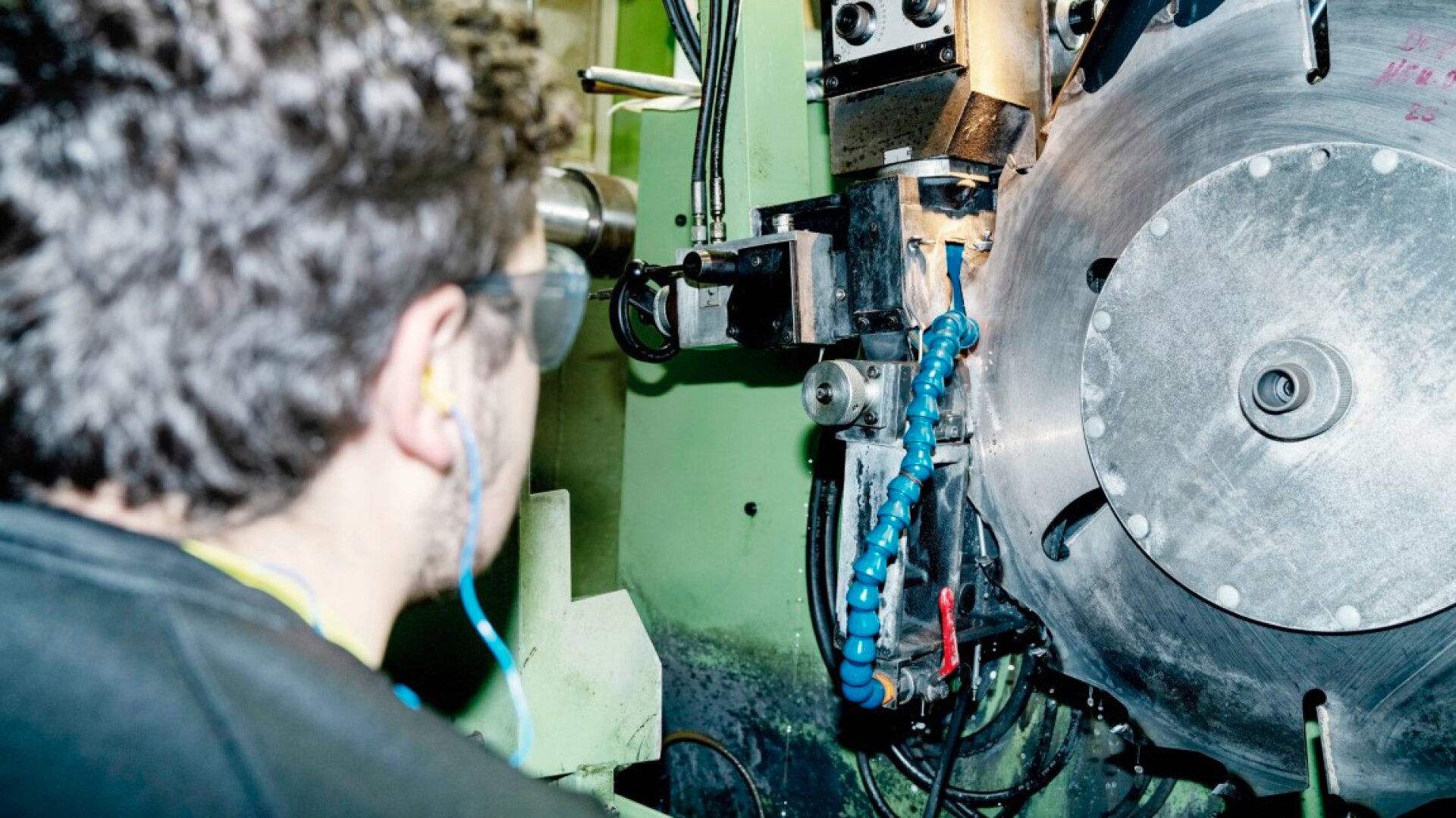 Eine Person, die eine Schutzbrille und einen Gehörschutz trägt, bedient oder überprüft in einer hell erleuchteten Werkstatt eine große Industriemaschine mit einem runden Metallteil und blauen flexiblen Schläuchen.