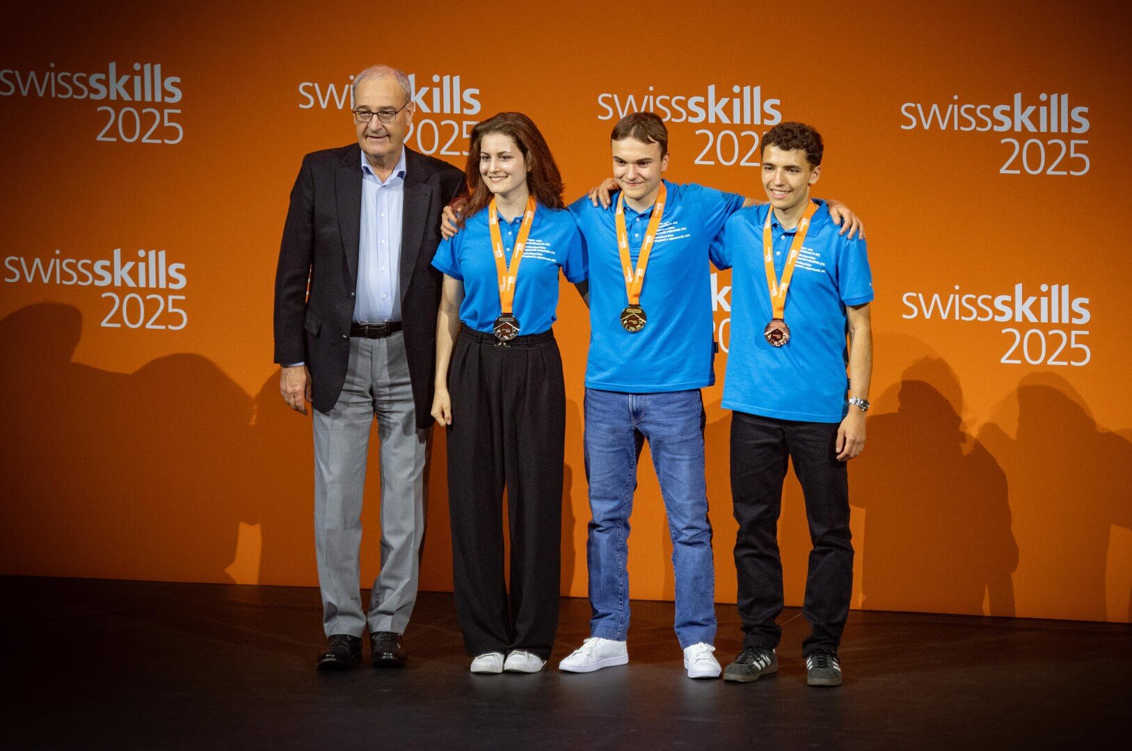 Vier Personen stehen vor einer orangefarbenen Kulisse der SwissSkills 2025. Drei junge Menschen in blauen Hemden tragen Medaillen und legen lächelnd die Arme umeinander, während ein älterer Mann im Anzug daneben steht.