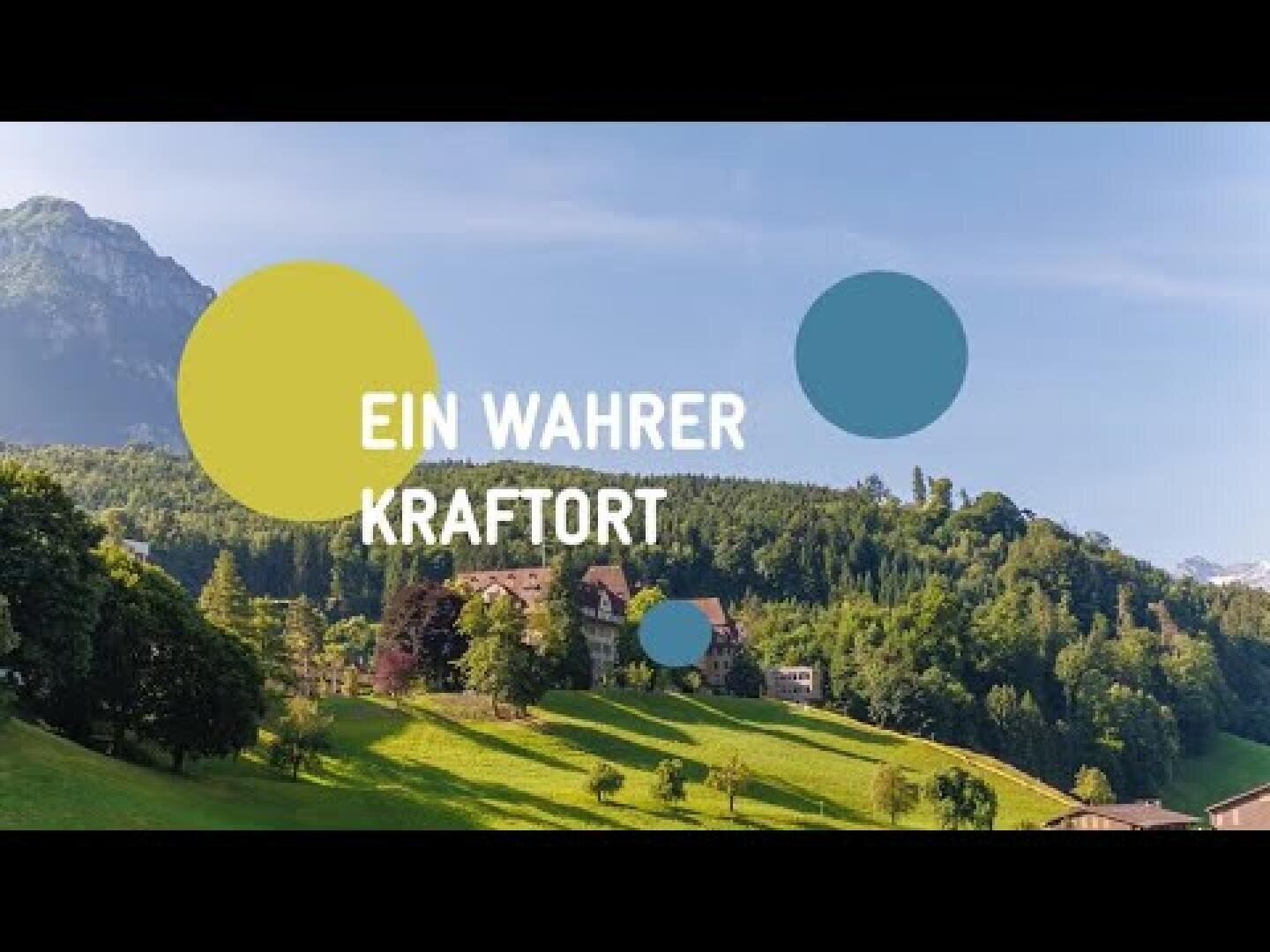 Eine malerische Ansicht eines üppigen grünen Hügels mit Bäumen und ein paar Gebäuden, im Hintergrund Berge unter einem blauen Himmel. Der überlagerte Text lautet EIN WAHRER KRAFTORT und ist von farbigen Kreisen umgeben.