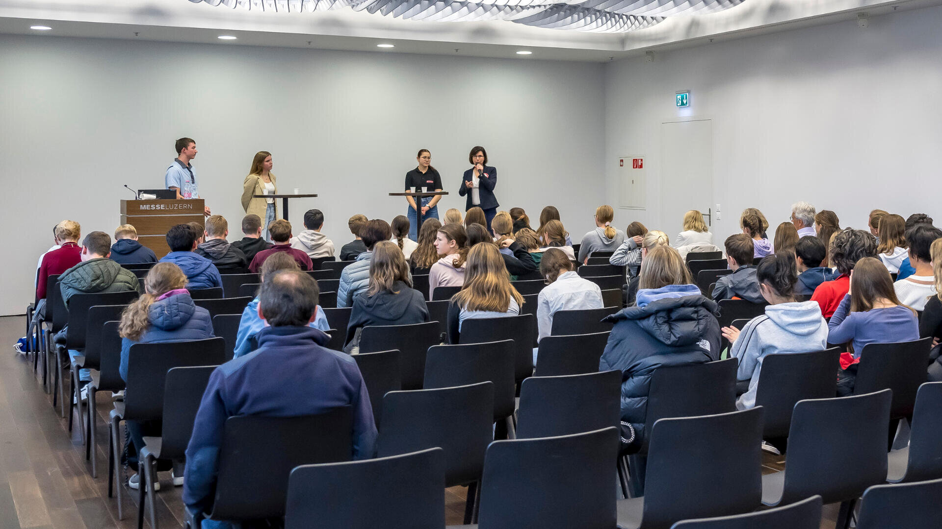 Eine Gruppe von Personen sitzt in Reihen vor einer Bühne, auf der vier Redner an Podien stehen und sich in einem modernen Konferenzraum an das Publikum wenden.