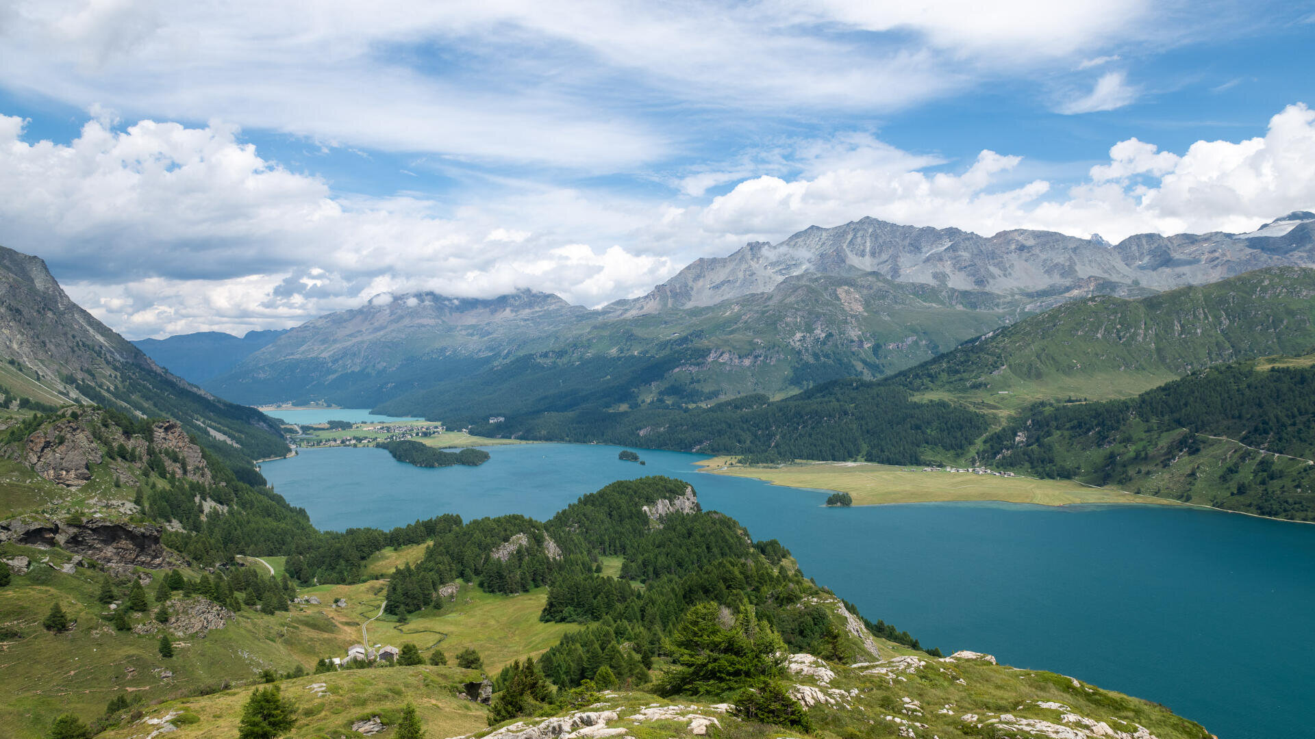 Ein Panoramablick auf einen blauen Alpensee, umgeben von üppig grünen Hügeln, vereinzelten Bäumen und schroffen Bergen unter einem teilweise bewölkten Himmel.