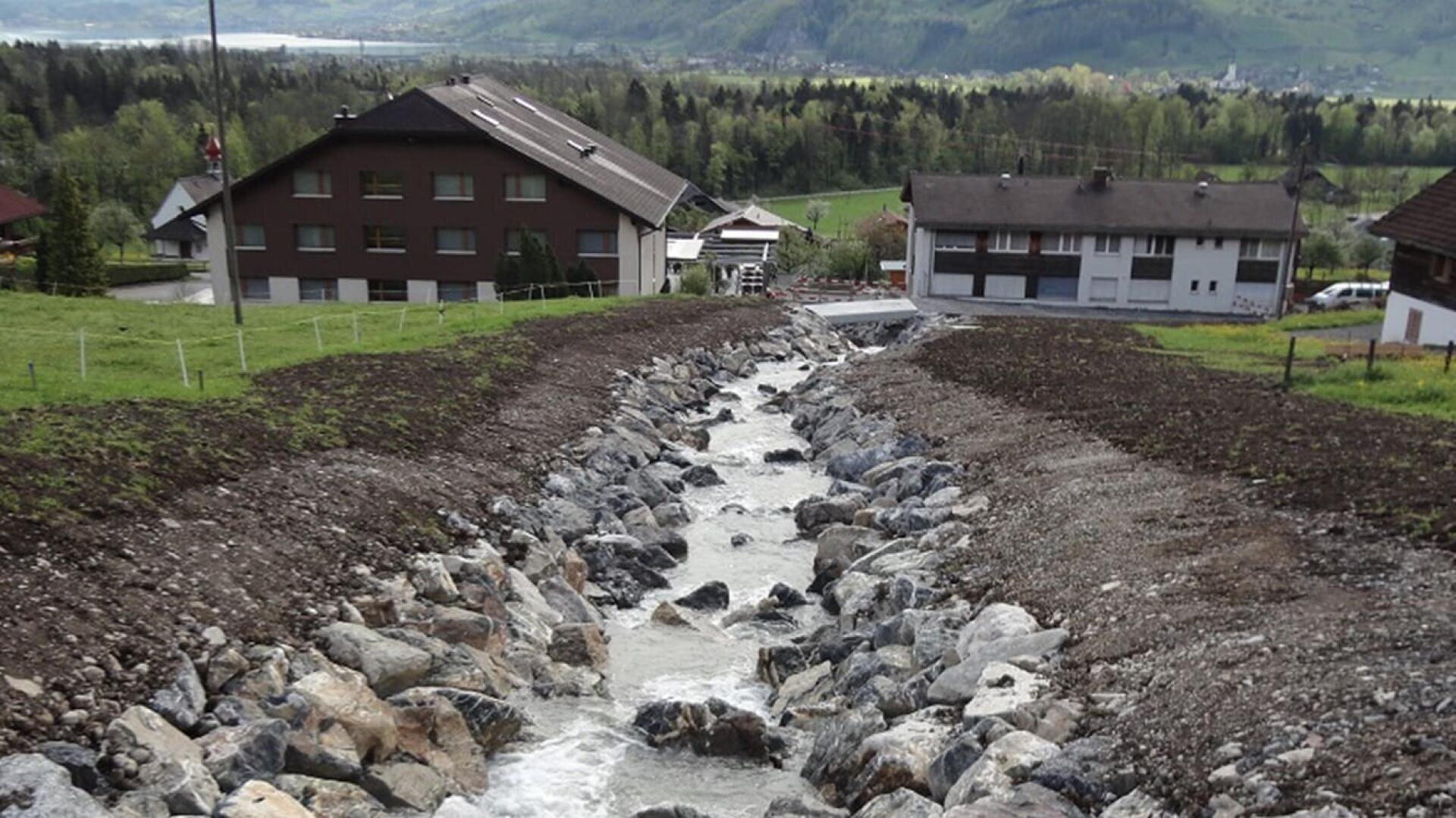 Ein felsiger Bach fließt zwischen grasbewachsenen Feldern bergab in Richtung der Häuser, mit grünen Hügeln und Bäumen im Hintergrund unter einem bewölkten Himmel.