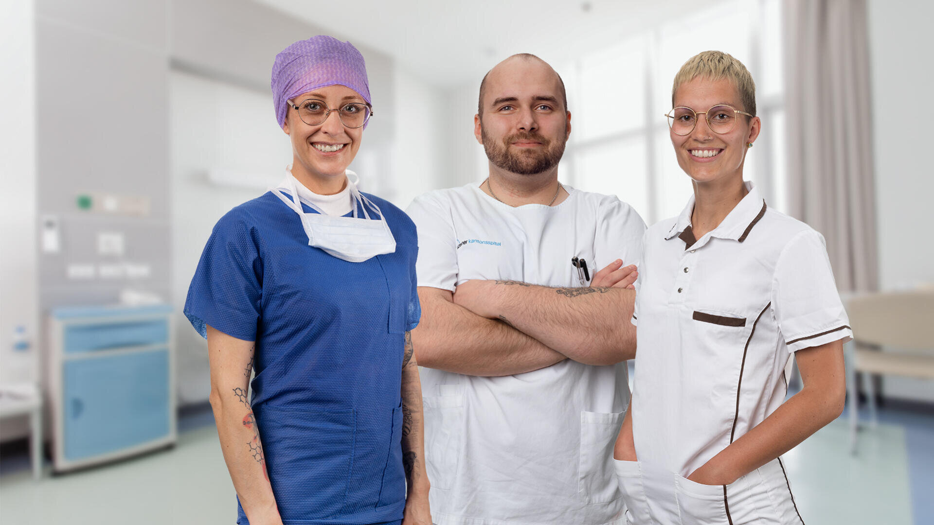 Drei medizinische Fachkräfte posieren und lächeln in einem Krankenhauszimmer. Zwei von ihnen tragen Brillen und medizinische Kittel - einer in blauem Kittel mit Kappe und Maske, der andere in weißem Kittel mit dunkler Verzierung. Der dritte, in weißem Kittel, steht mit verschränkten Armen da.
