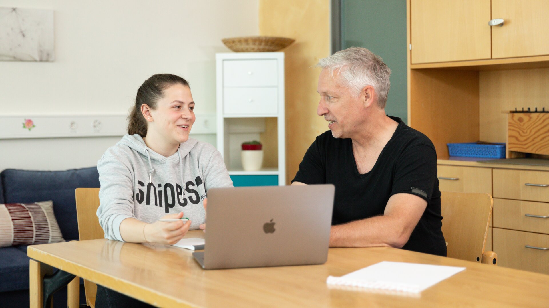 Eine jüngere Frau und ein älterer Mann sitzen an einem Tisch in einem legeren Raum, lächeln und unterhalten sich. Ein Laptop, ein Notizblock und ein Stift liegen auf dem Tisch zwischen ihnen. Im Hintergrund sind Regale und ein Sofa zu sehen.
