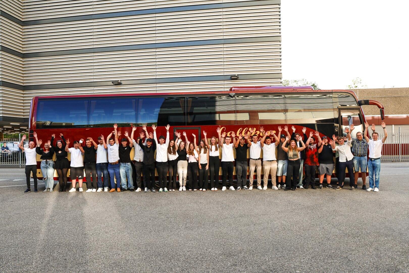 Eine Gruppe von Menschen steht in einer Reihe vor einem roten Tourbus, lächelt und hebt feierlich die Arme. Im Hintergrund sind moderne Gebäude und Zäune zu sehen.