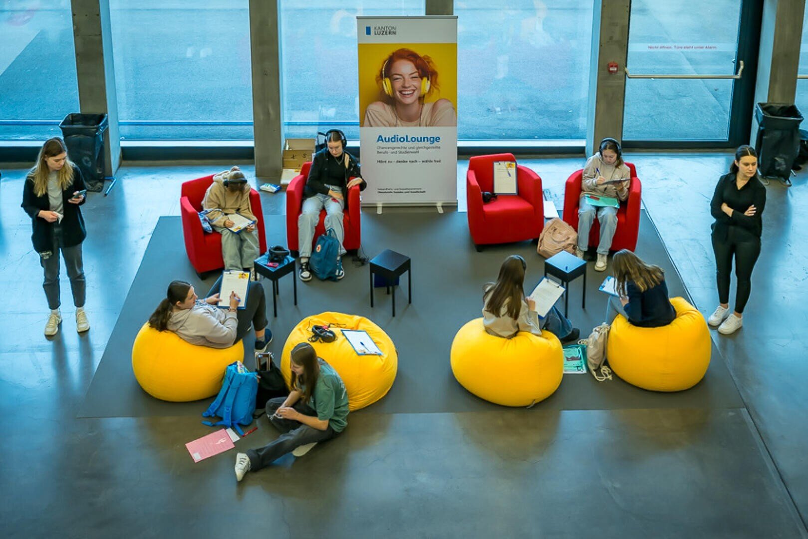 Studierende sitzen auf gelben Sitzsäcken und roten Stühlen in einem modernen Lounge-Bereich mit großen Fenstern, lesen oder arbeiten an Laptops. In der Mitte steht ein Banner mit einer lächelnden Person und dem Text "AudioLounge".