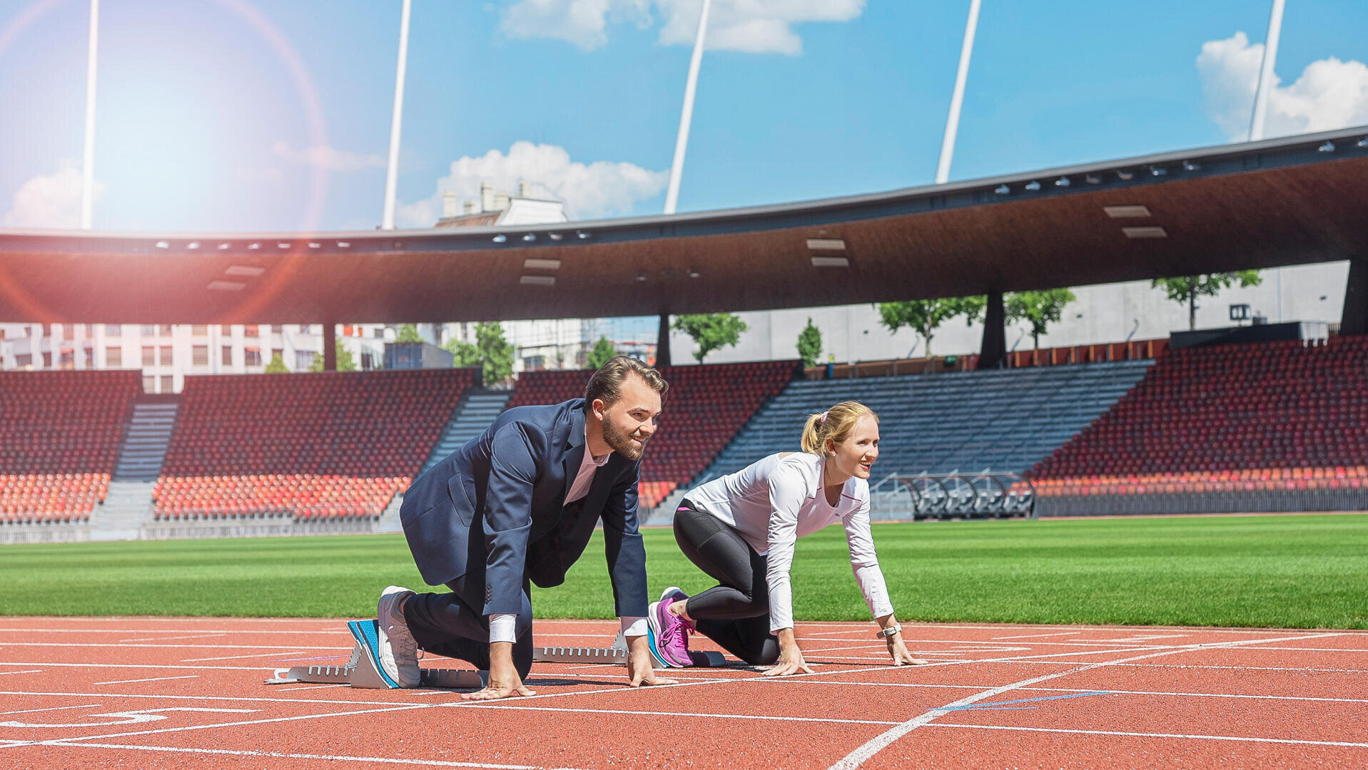 Ein Mann im Geschäftsanzug und eine Frau in Sportkleidung kauern an der Startlinie auf einer Laufbahn in einem Stadion, bereit zum Rennen, mit leeren roten Sitzen und einem blauen Himmel im Hintergrund.