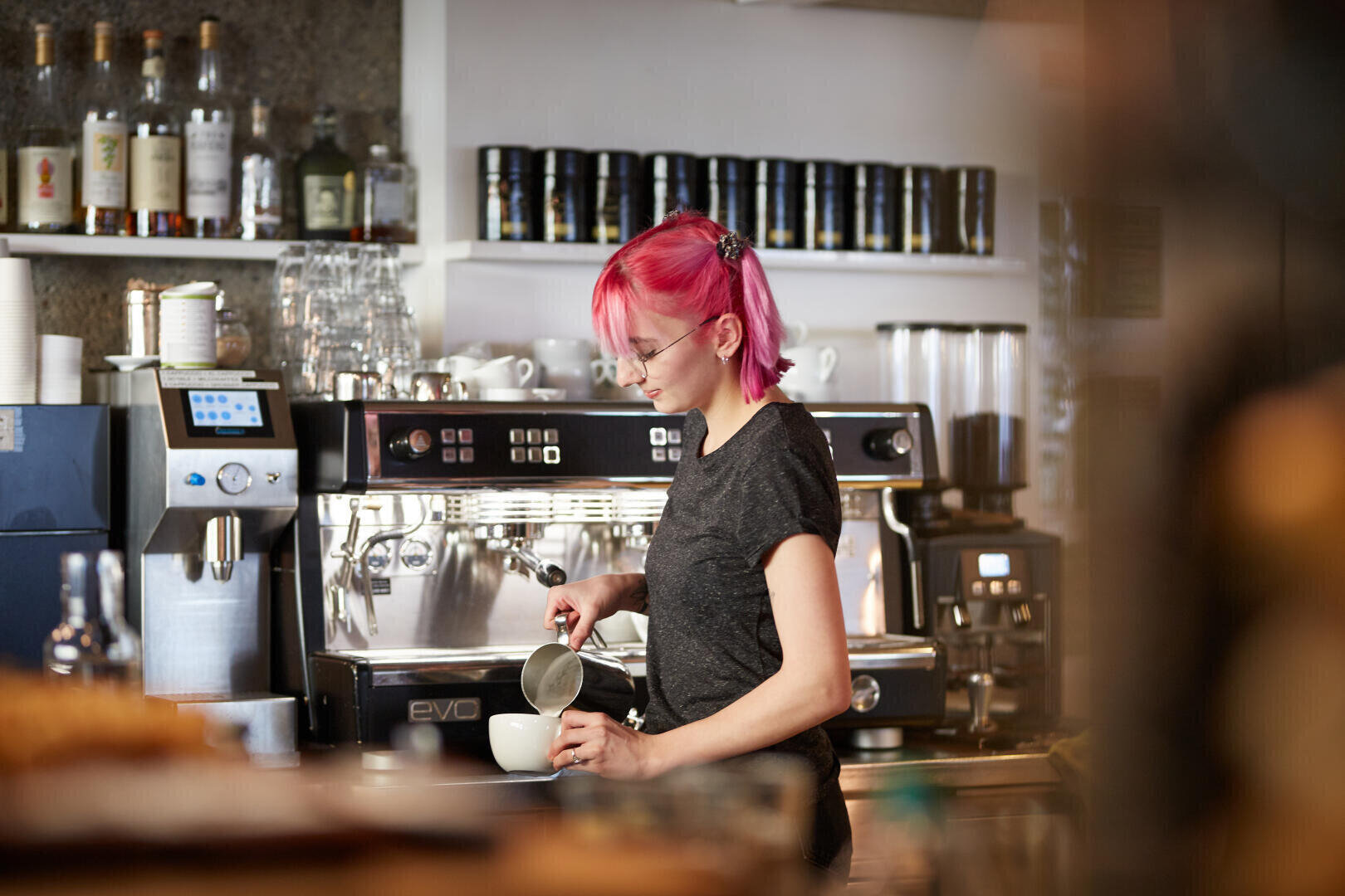 Ein Barista mit rosa Haaren und Brille brüht Milch an einer Espressomaschine in einem modernen Coffeeshop auf, während im Hintergrund Flaschen und Tassen in Regalen stehen.