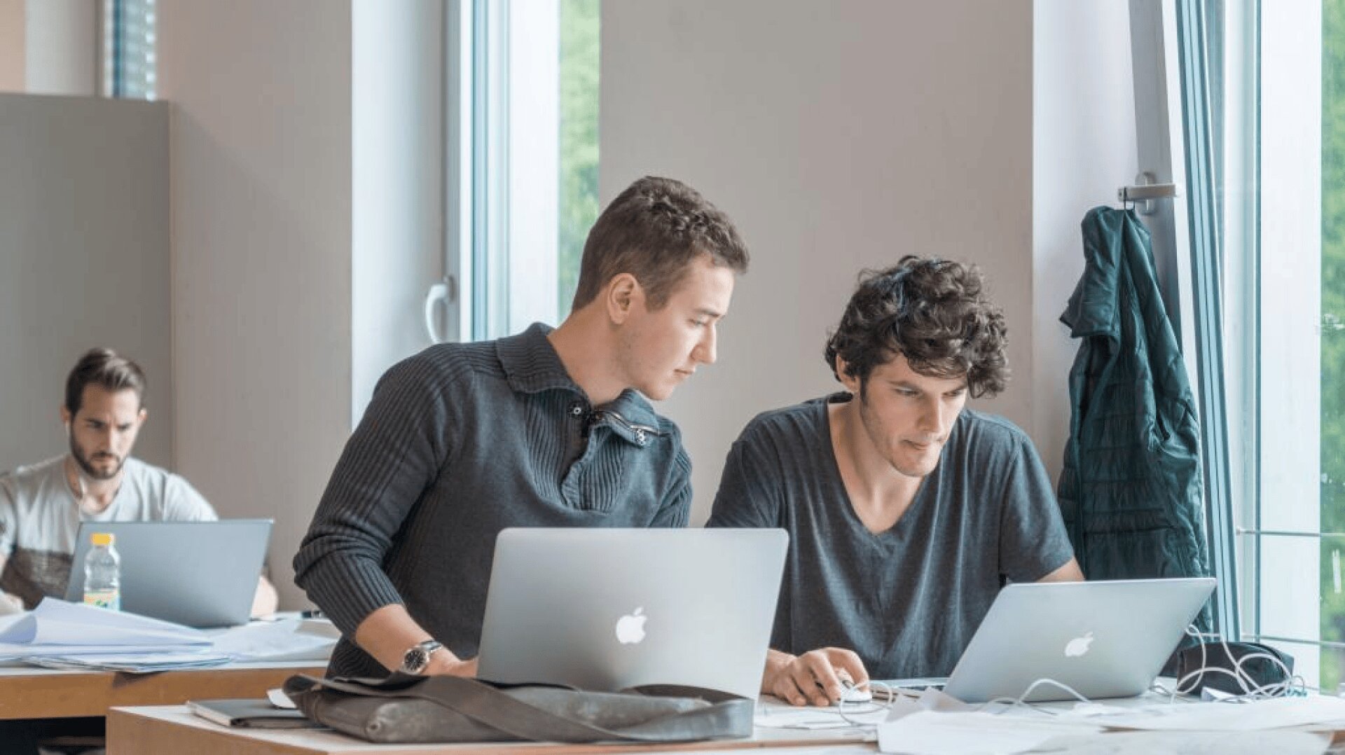 Zwei Männer sitzen an einem Tisch und arbeiten an Laptops, wobei einer steht und sich vorbeugt, um dem anderen zu helfen. Im Hintergrund sitzt eine weitere Person, die ebenfalls an einem Laptop arbeitet. Auf dem Tisch liegen Papiere und Flaschen.