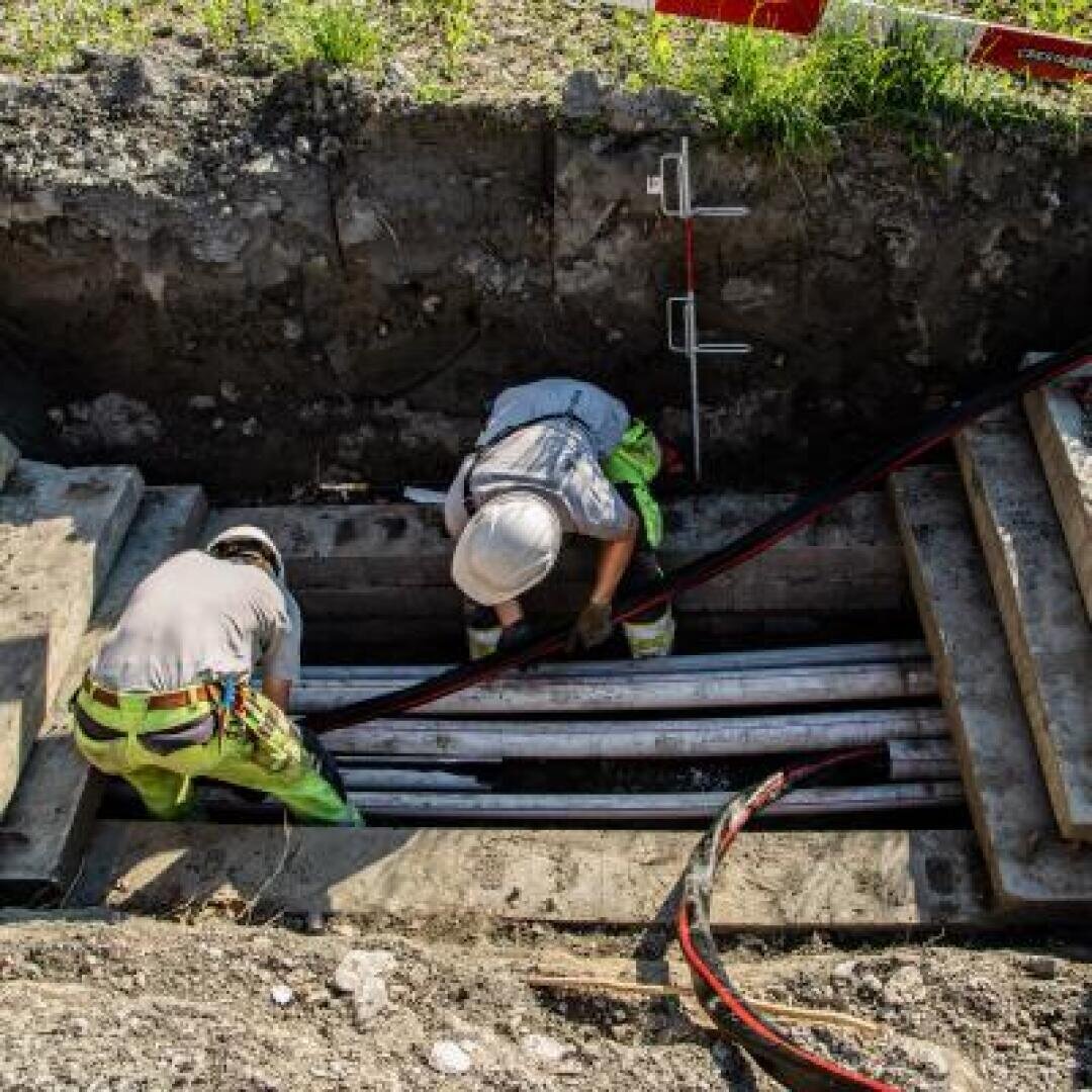Zwei Bauarbeiter in Schutzkleidung arbeiten in einem Graben und verlegen Rohre und Kabel. Der Graben ist von Erde umgeben, und am Rand sind Baumaschinen und eine Absperrung zu sehen.