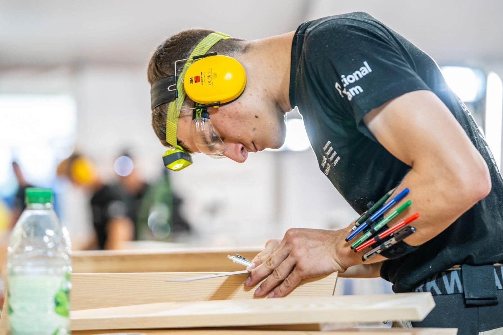 Ein junger Mann, der einen Gehörschutz, eine Schutzbrille und eine Stirnlampe trägt, ist mit Holzarbeiten beschäftigt. Er schreibt auf einem Holzbrett mit mehreren Stiften, die an seinem Arm befestigt sind. Auf dem Tisch daneben steht eine Plastikflasche.