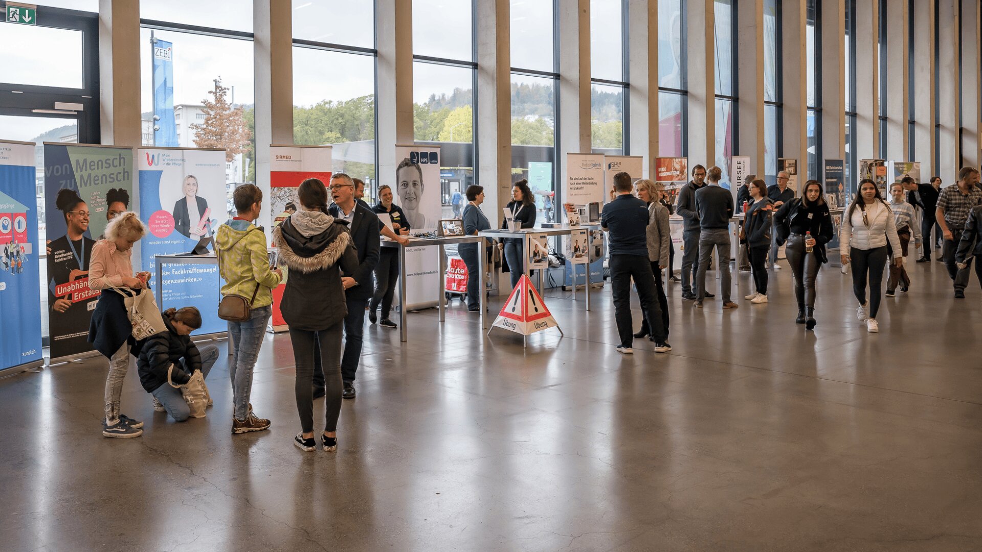 Die Menschen gehen und unterhalten sich in einer geräumigen, modernen Halle mit großen Fenstern und mehreren Informationsständen an den Seiten. Einige Erwachsene unterhalten sich mit Kindern, während andere die Auslagen durchstöbern oder sich in kleinen Gruppen unterhalten.