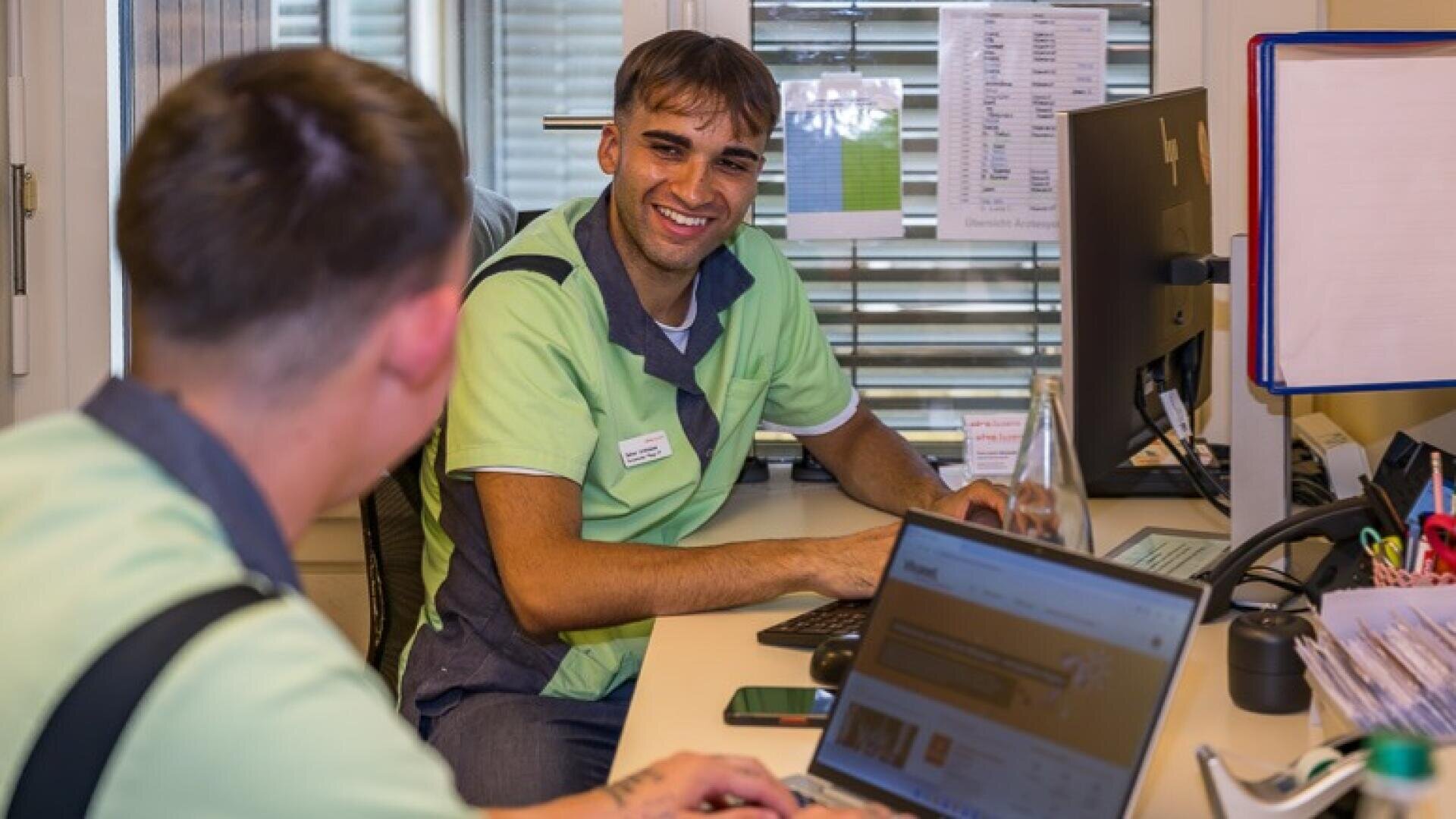 Ein lächelnder Mitarbeiter im Gesundheitswesen in grüner Uniform sitzt an einem Schreibtisch und unterhält sich mit einem Kollegen, während er am Computer arbeitet.