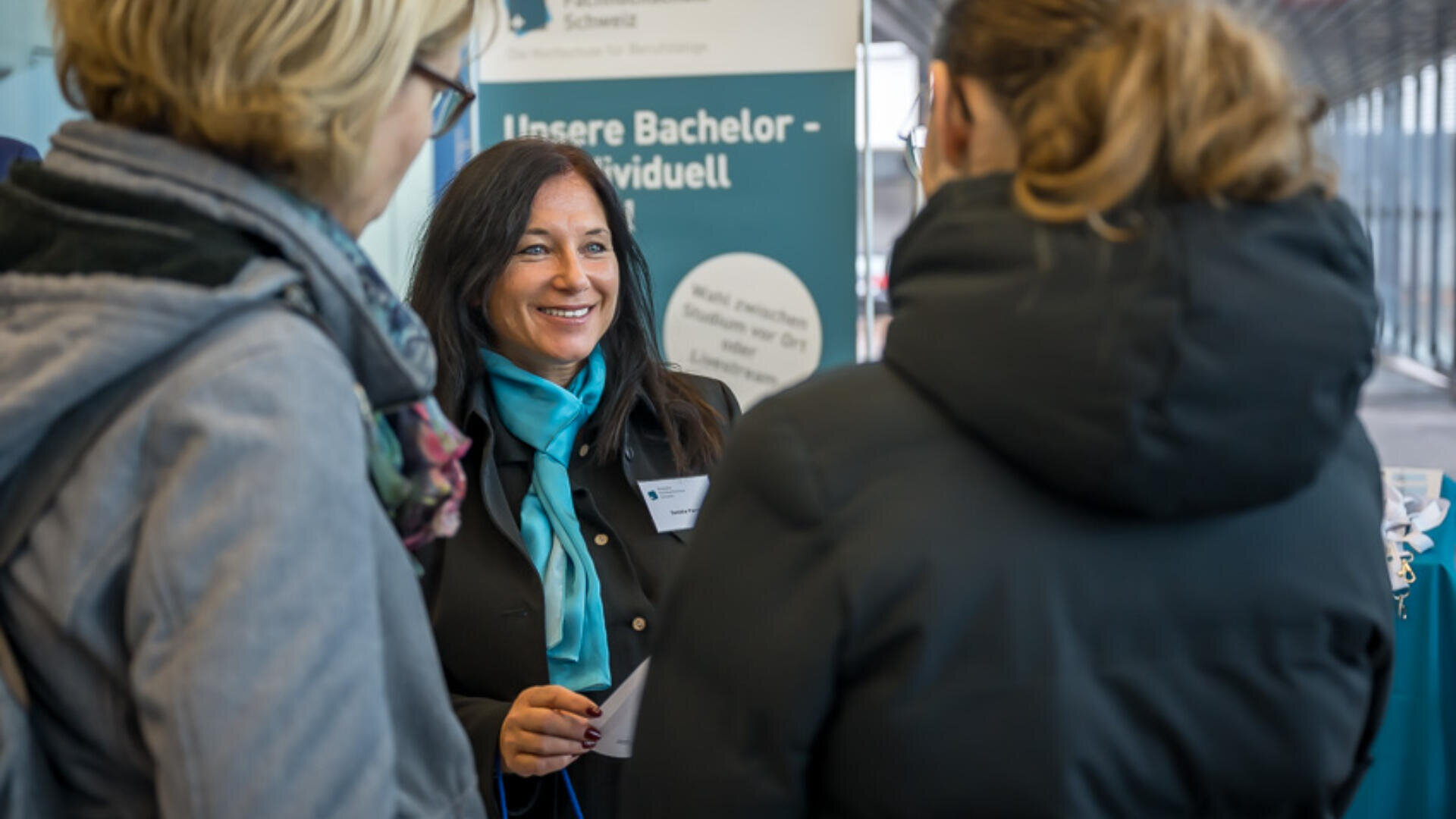 Eine lächelnde Frau mit einem türkisfarbenen Schal unterhält sich mit zwei Personen an einem Informationsstand. Im Hintergrund ist ein Schild mit Informationen über Bachelor-Studiengänge in deutscher Sprache zu sehen.