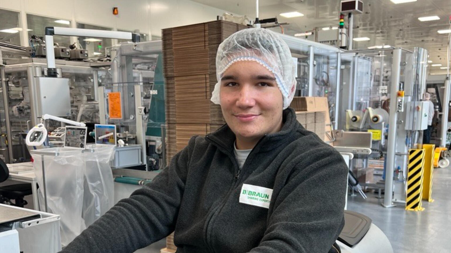 Junge Person, die ein Haarnetz und eine dunkle Jacke mit einem Namensschild trägt und lächelt, während sie in einer Produktions- oder Verpackungsanlage mit Maschinen und Kartons im Hintergrund sitzt.