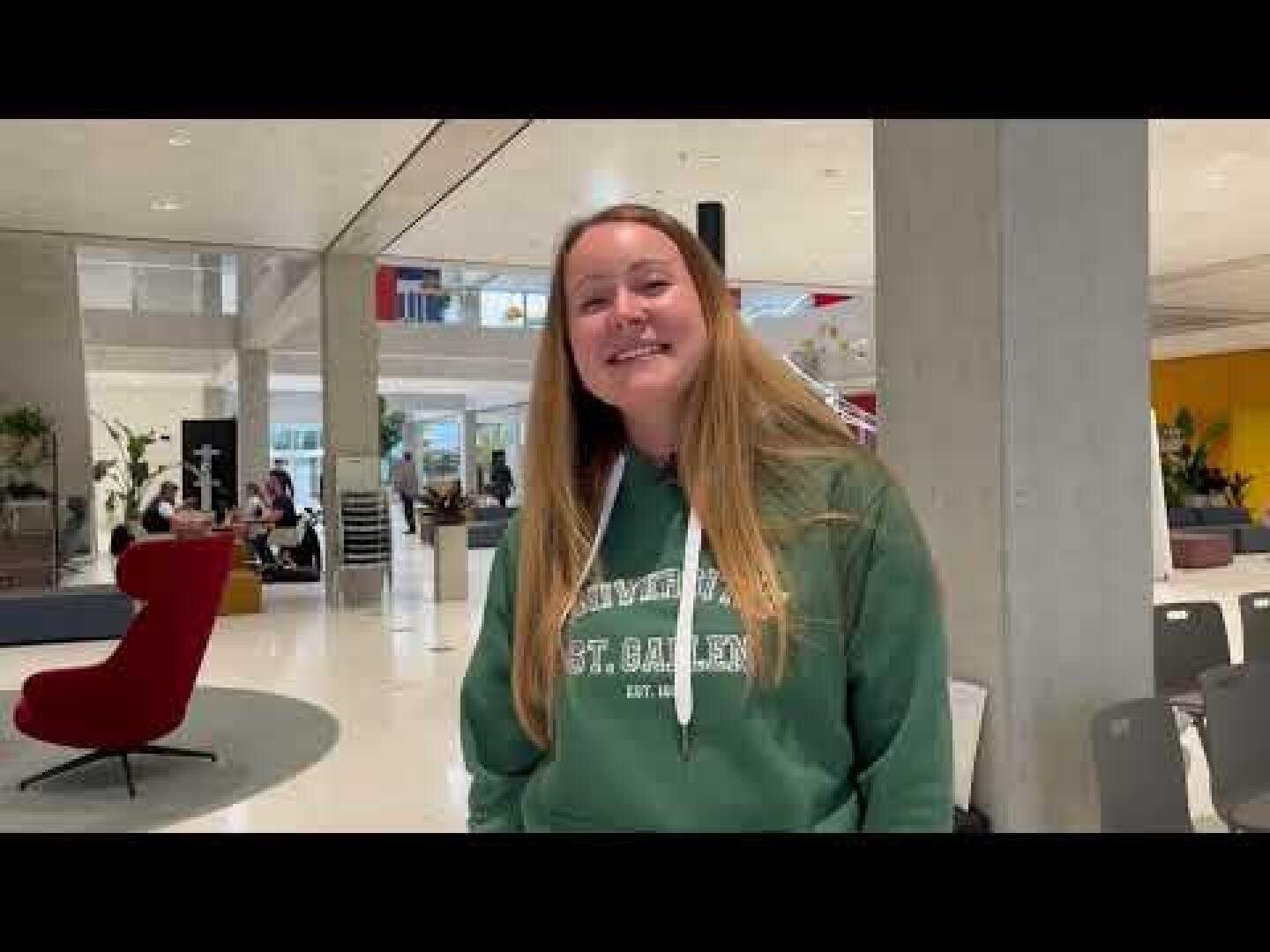 Eine Frau mit langen blonden Haaren, die einen grünen Kapuzenpulli der Universität St. Gallen trägt, lächelt, während sie in einem modernen, offenen Innenraum mit Stühlen, Tischen und mehreren Personen im Hintergrund steht.