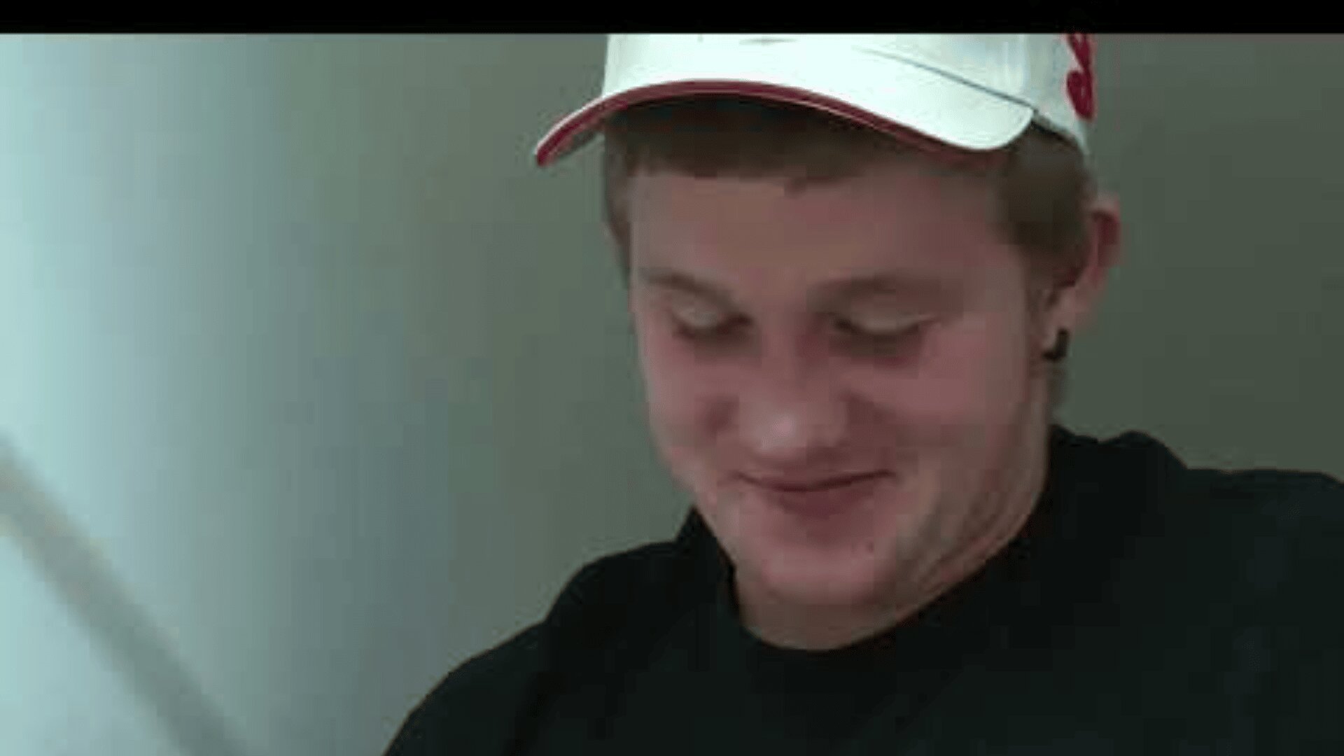 Ein junger Mensch mit weißer Mütze und schwarzem Hemd blickt nach unten und lächelt sanft. Der Hintergrund ist schlicht und in hellen Farben gehalten.