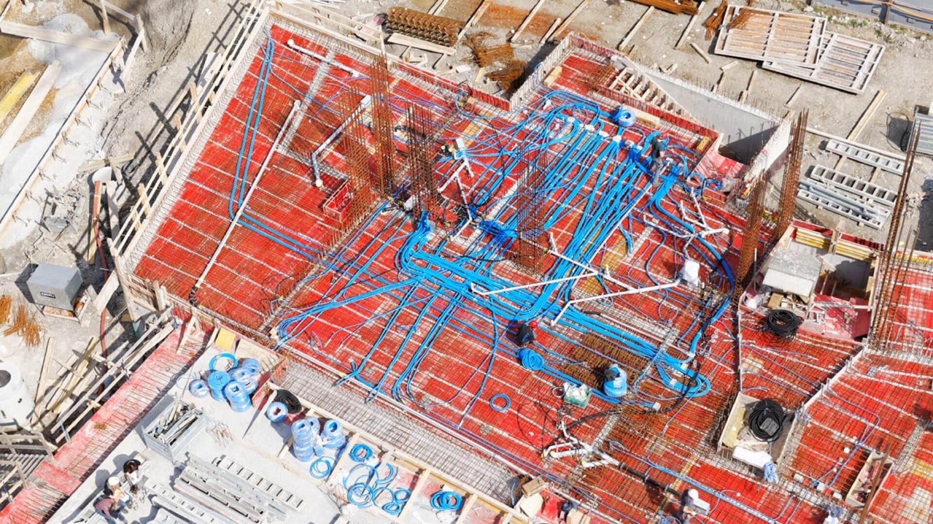 Luftaufnahme einer Baustelle, die ein Gebäudefundament mit freiliegendem Bewehrungsstahl, blauen Elektro- oder Sanitärleitungen und verstreutem Baumaterial zeigt. Arbeiter sind auf der Baustelle anwesend.