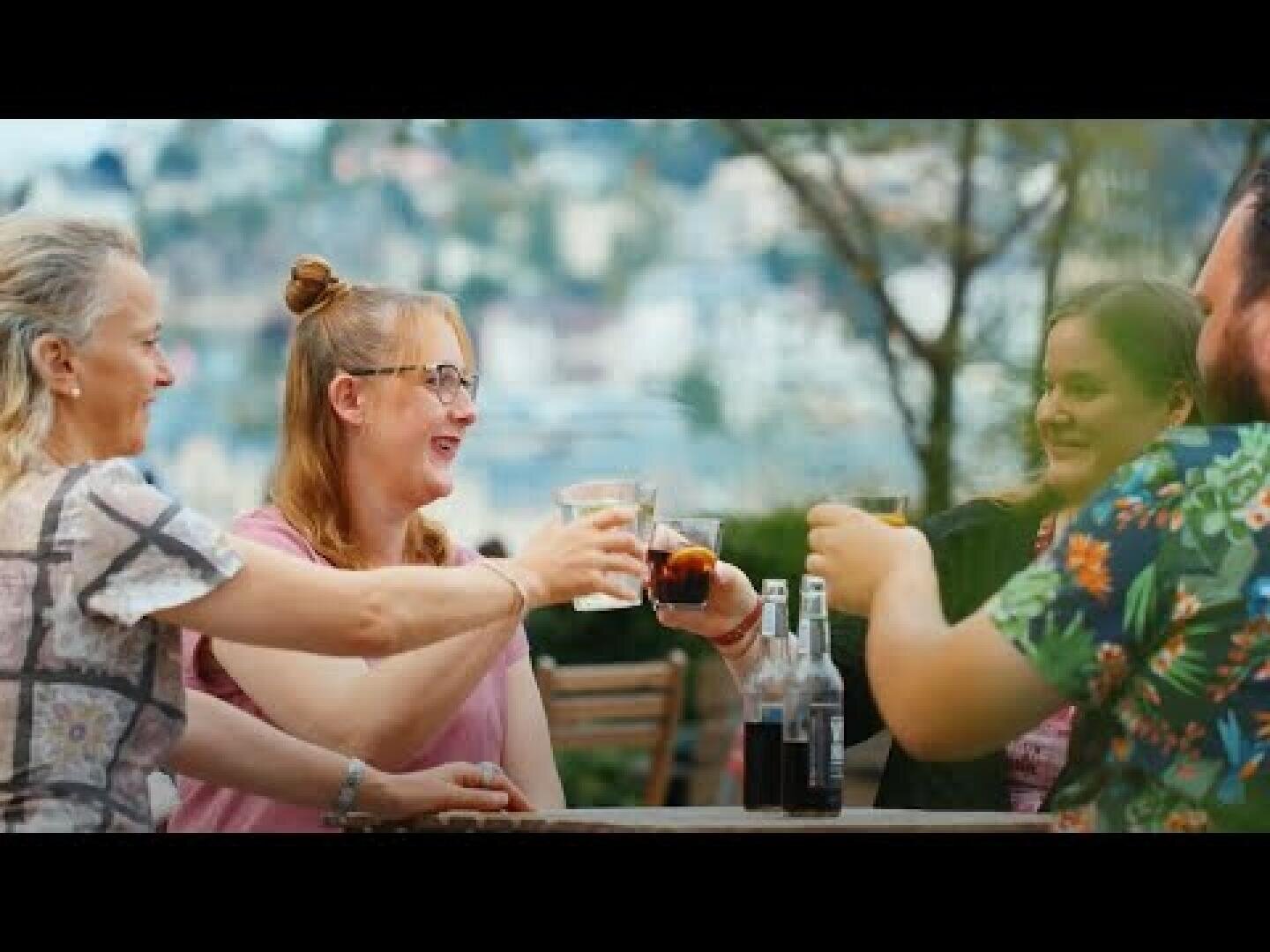 Vier Personen sitzen an einem Tisch im Freien, lächeln und stoßen mit ihren Gläsern an. Der Hintergrund ist unscharf und zeigt Bäume und eine Stadtlandschaft. Auf dem Tisch stehen Flaschen und Getränke.