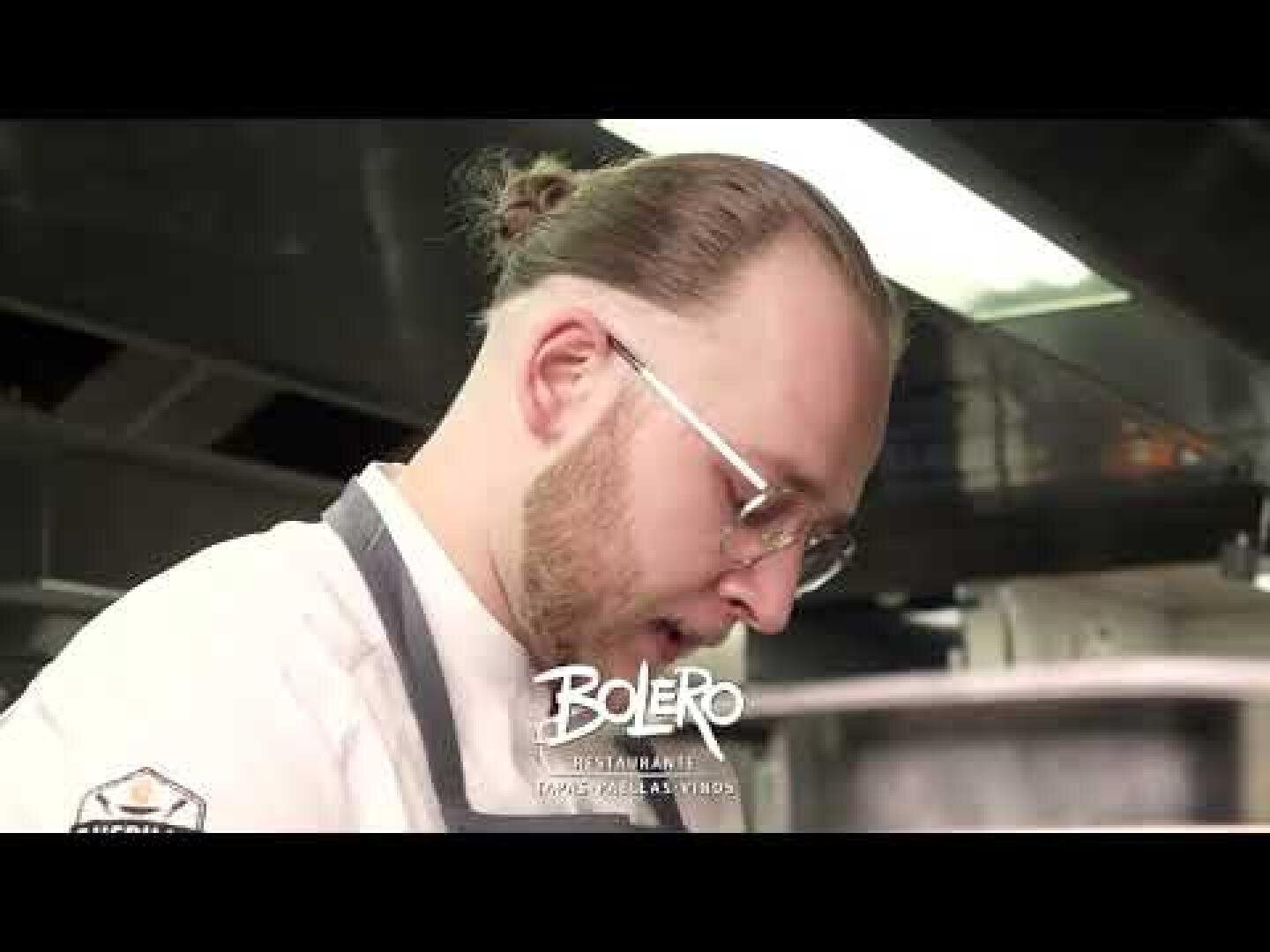 Ein Koch mit Brille und Bart, der einen weißen Kittel und eine Schürze trägt, arbeitet in einer Küche. Im Hintergrund sind Flächen aus rostfreiem Stahl zu sehen. Am unteren Rand des Bildes steht "Bolero Restaurant" geschrieben.
