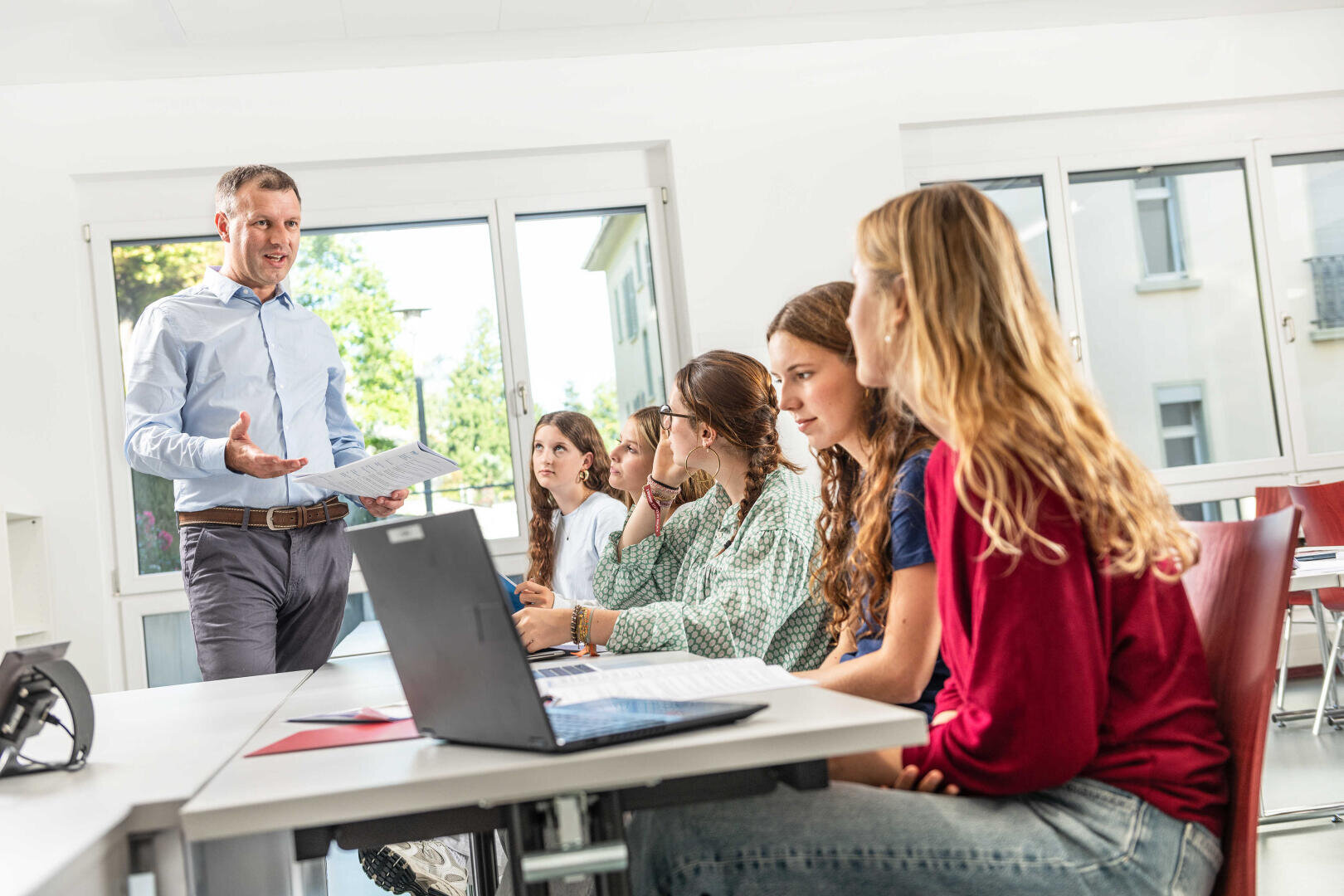 Ein Mann steht vorne in einem hellen Klassenzimmer und spricht zu einer Gruppe von vier Schülern, die mit Laptops und Notebooks an den Tischen sitzen, während hinter ihnen das Sonnenlicht durch große Fenster strömt.