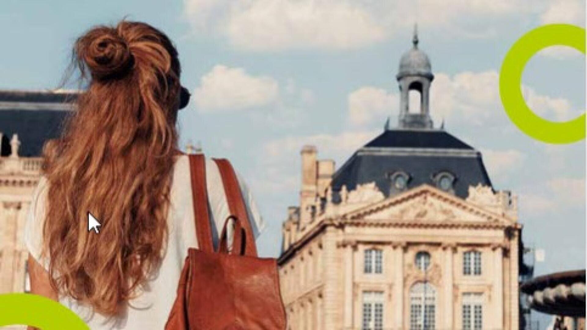 Eine Frau mit langen Haaren und einem braunen Rucksack steht unter bewölktem Himmel vor einem historischen Steingebäude mit einer Kuppel.