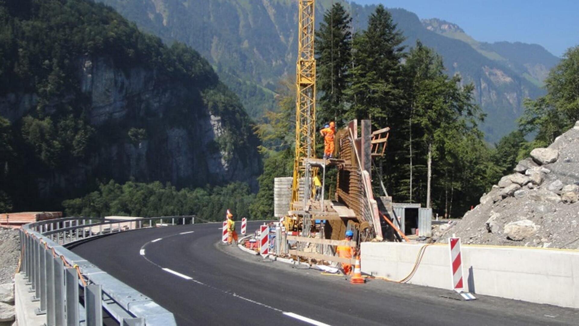 Arbeiter in orangefarbener Schutzkleidung errichten mit einem Gerüst und einem Kran ein Bauwerk am Straßenrand neben einer neu asphaltierten Straße, die sich durch eine bergige, bewaldete Landschaft schlängelt.