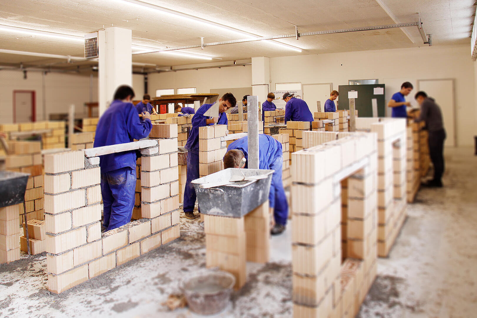 Mehrere Personen in blauen Arbeitsuniformen sind dabei, in einem Innenraum, möglicherweise in einer Werkstatt oder einem Schulungsraum, mit verschiedenen Werkzeugen und Materialien Mauern zu errichten.