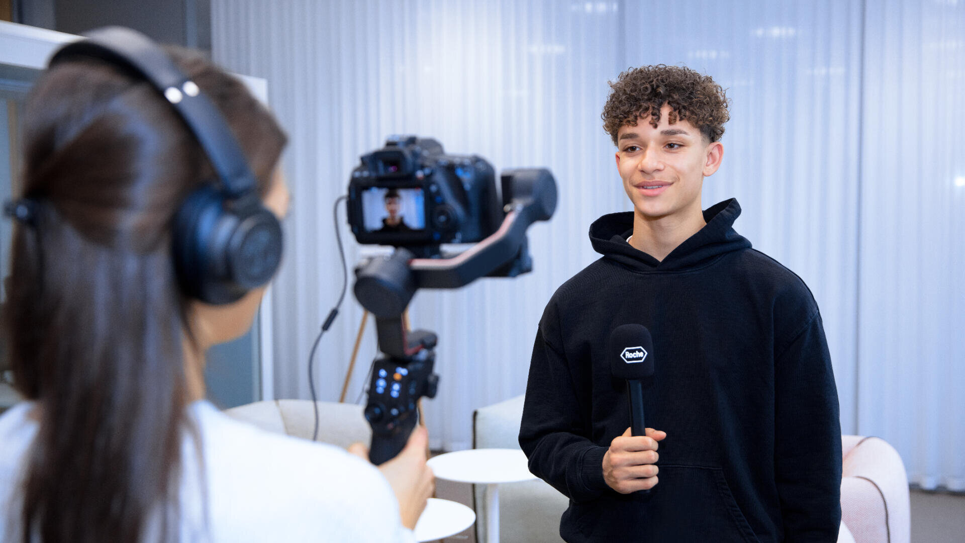 Eine junge Person in einem schwarzen Kapuzenpulli hält ein Mikrofon in der Hand und spricht, während sie von einer Person gefilmt wird, die Kopfhörer trägt und eine kardanisch aufgehängte Kamera in einer modernen Innenumgebung bedient.