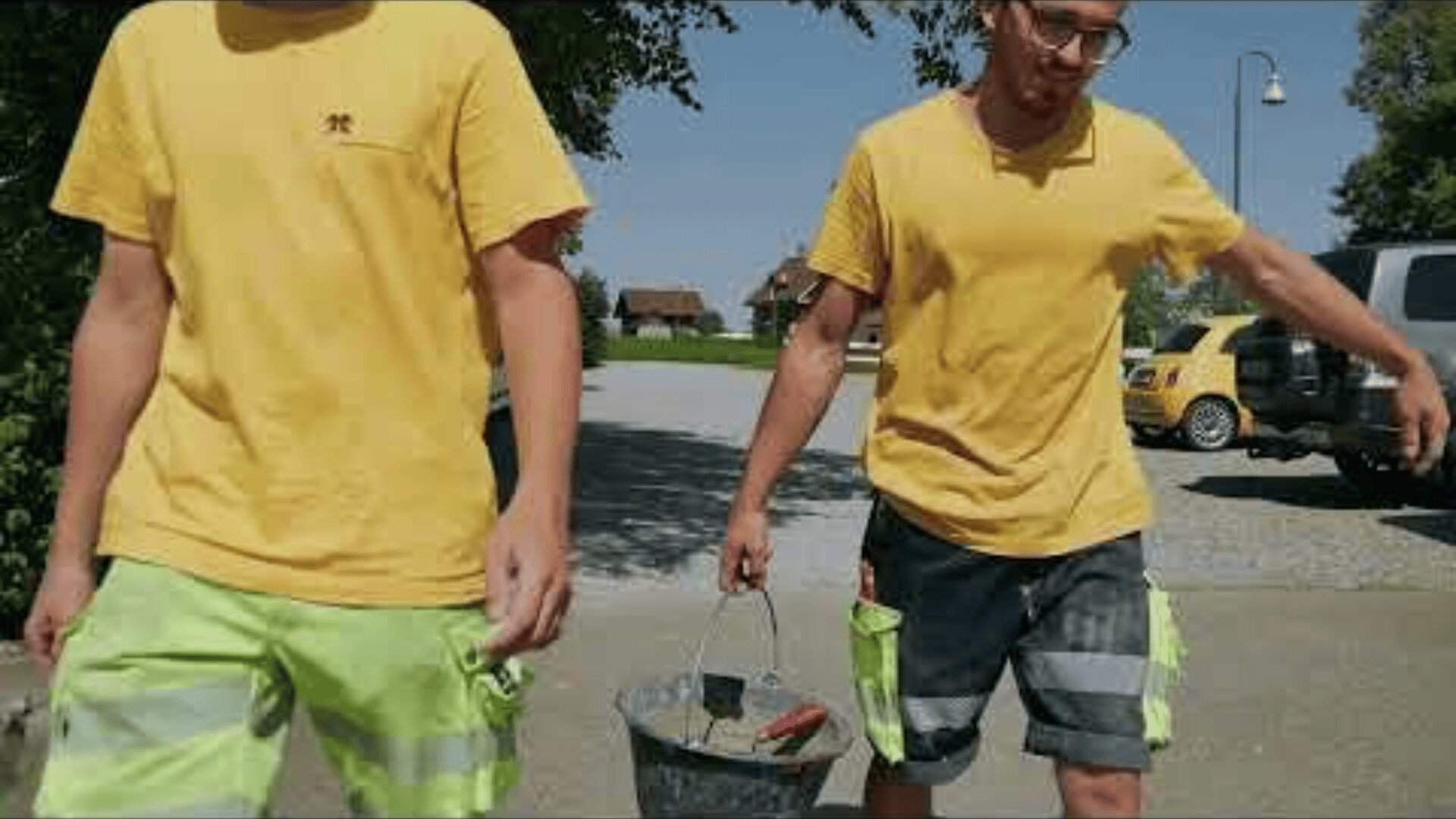Zwei Personen in gelben Hemden und Arbeitsshorts gehen an einem sonnigen Tag im Freien spazieren; eine trägt einen mit Werkzeugen gefüllten Eimer. Im Hintergrund sind ein gelbes Fahrzeug und Bäume zu sehen.