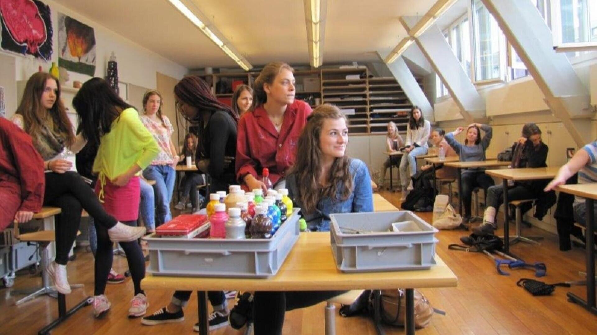 Ein lebhaftes Klassenzimmer mit Schülern, die stehen, sitzen und sich unterhalten. Auf einem Schreibtisch im Vordergrund liegen Malutensilien, und durch die großen Fenster auf der rechten Seite des Raums fällt Sonnenlicht.