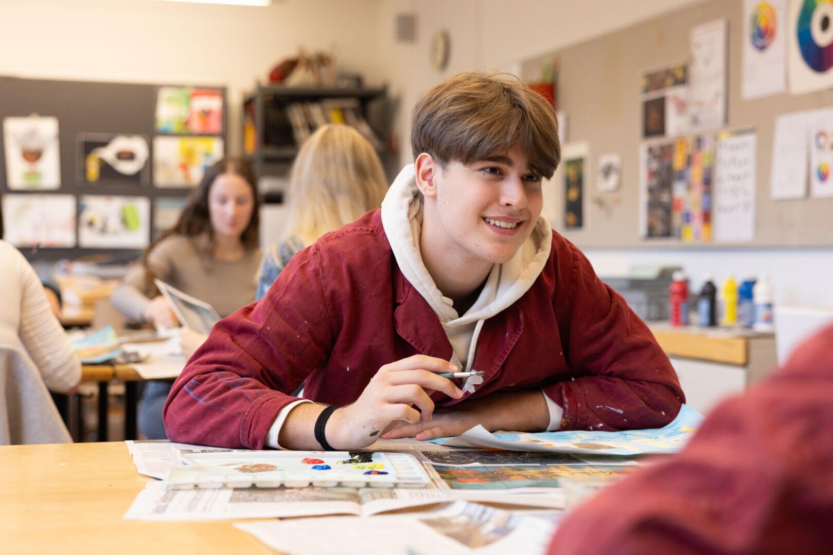 Ein lächelnder Schüler in einem roten Kittel sitzt an einem Tisch in einem Kunstunterrichtsraum, hält einen Pinsel und arbeitet an einem Bild. Im Hintergrund sind andere Schüler und Kunstzubehör zu sehen.